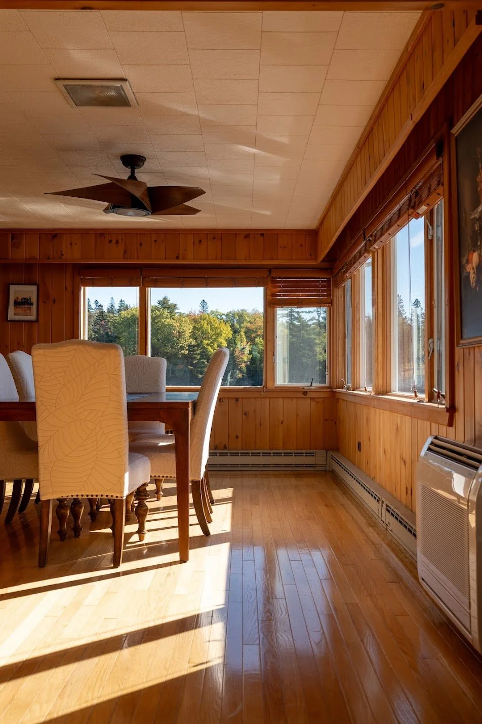 Sunlit dining room with wooden walls, hardwood floor, white upholstered chairs around a wooden table, large windows showcasing a green outdoor landscape, ceiling fan, and wall heater.