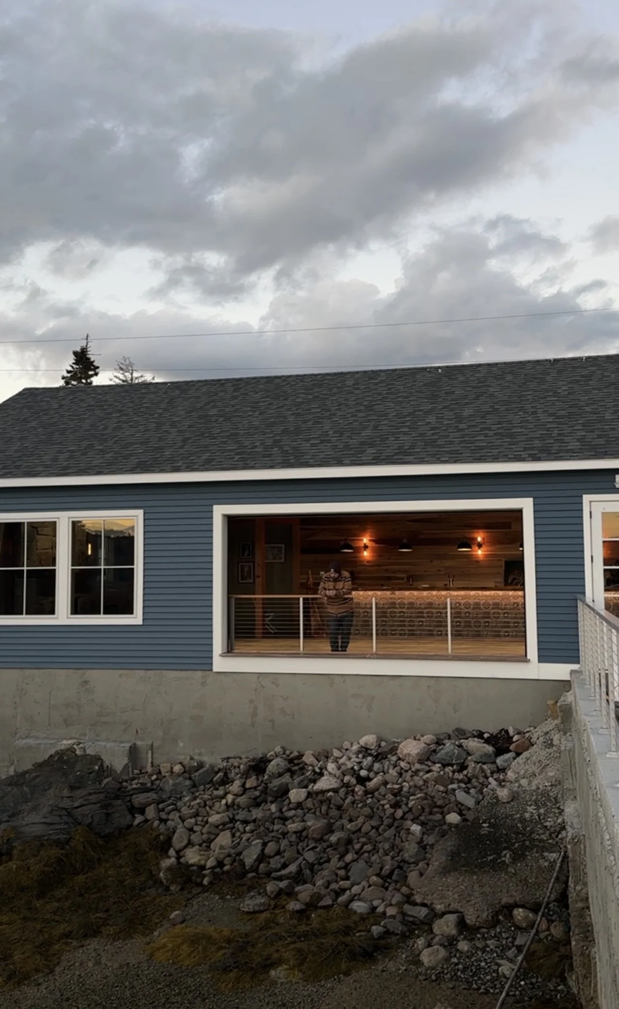 A person standing on a balcony of a blue house with a stone foundation, overlooking a rocky yard, with a cloudy sky overhead during dusk.