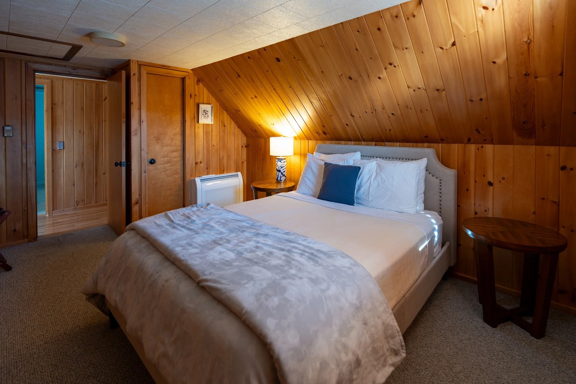 A cozy bedroom with wood-paneled walls and ceiling, featuring a neatly made bed with white and blue pillows, a bedside table with a lamp, and a closed door.