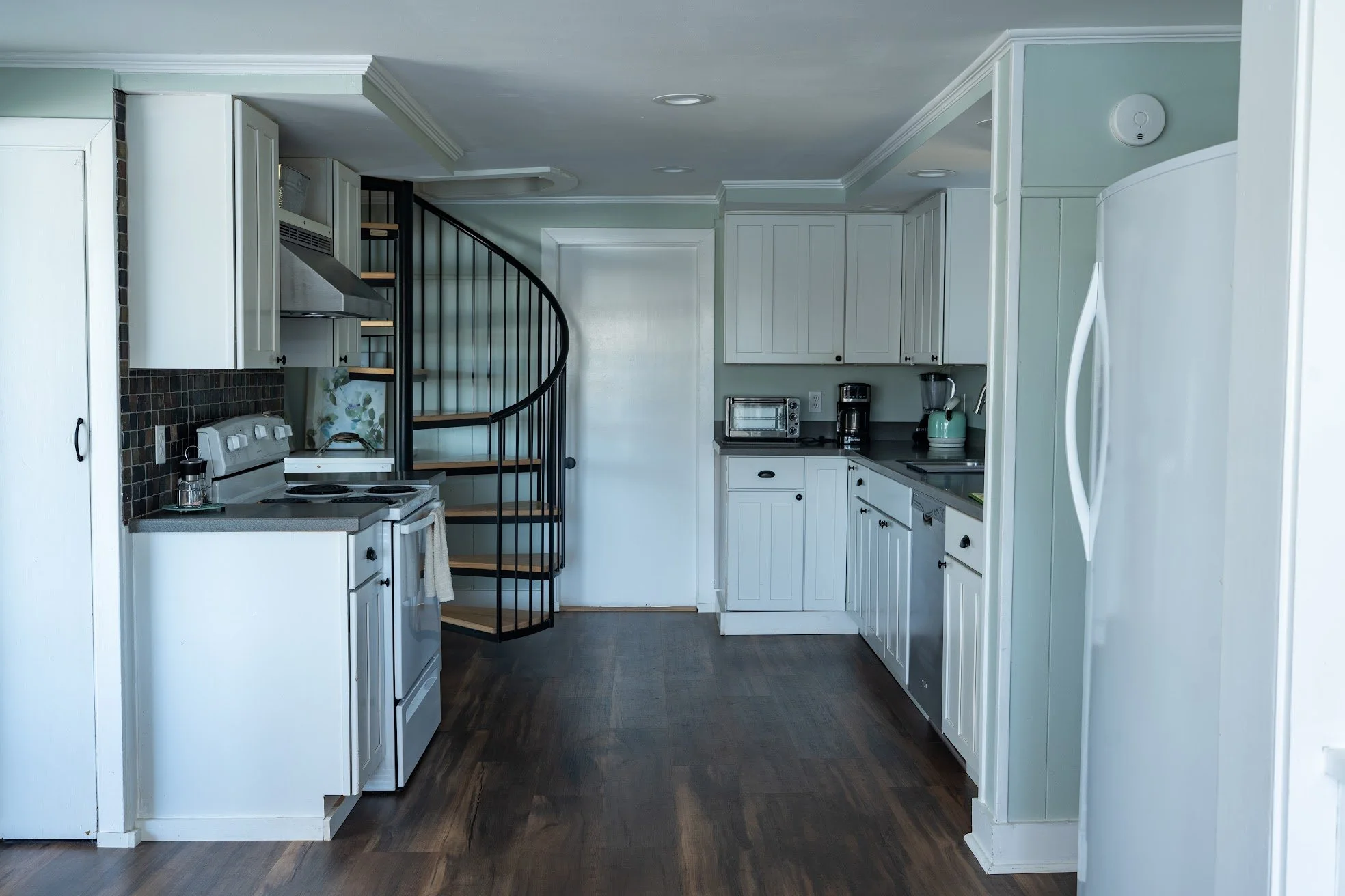 Modern kitchen with white cabinets, dark countertops, and wooden flooring. Includes appliances like a stove, microwave, toaster, coffee maker, and refrigerator. Spiral staircase with black railing in the background.