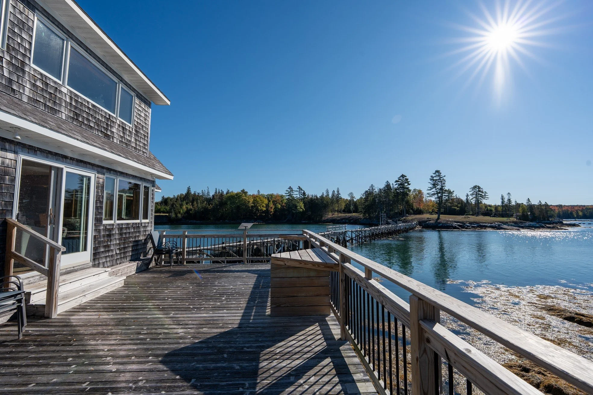 A wooden deck extends from a house overlooking a calm body of water with a small pier in the background. The house has large windows and weathered wooden siding. The sky is clear and bright with the sun shining.