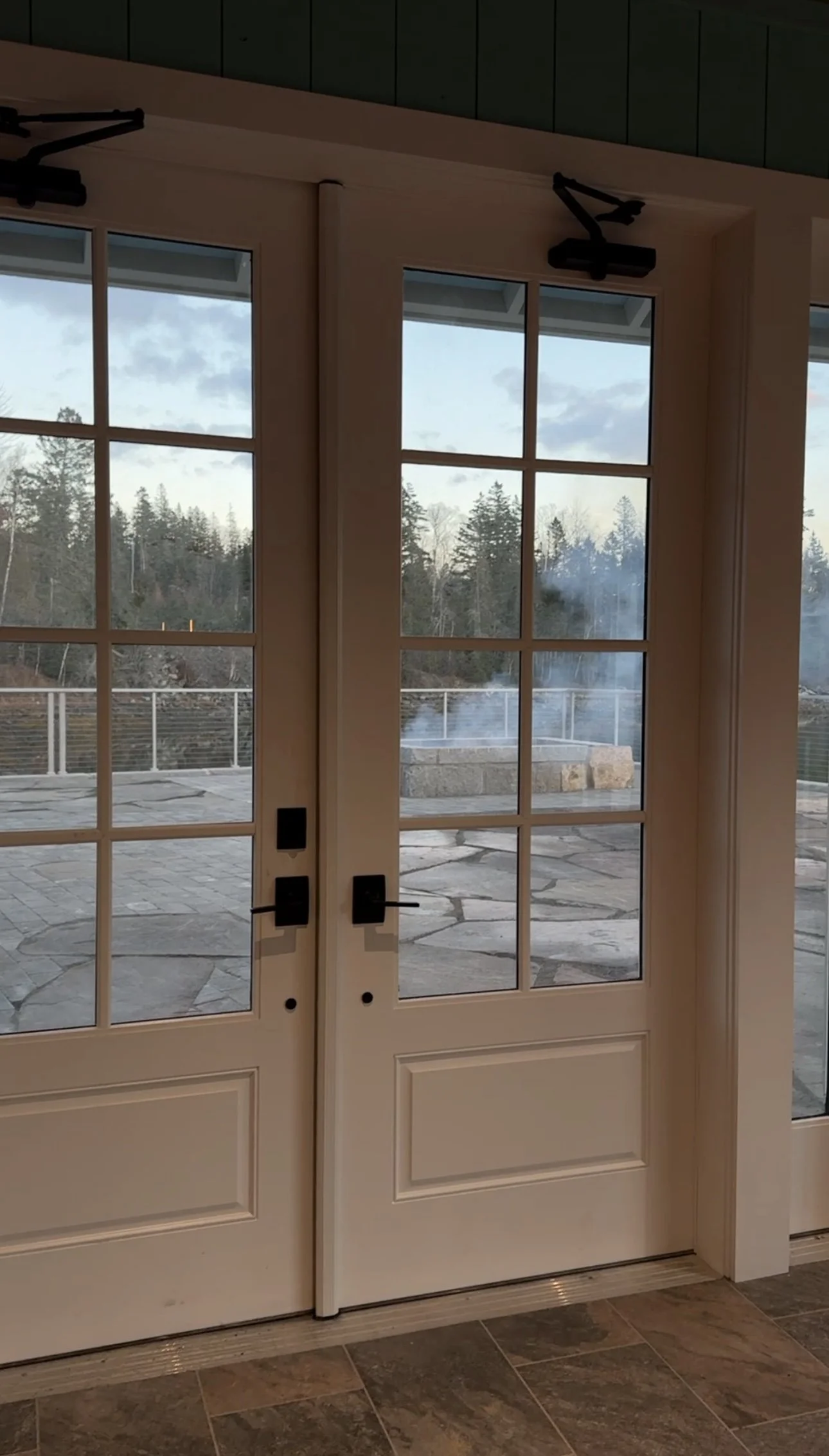 Interior view of double glass doors with black hardware, leading to an outdoor patio with stone paving and scenic forested landscape in the background.