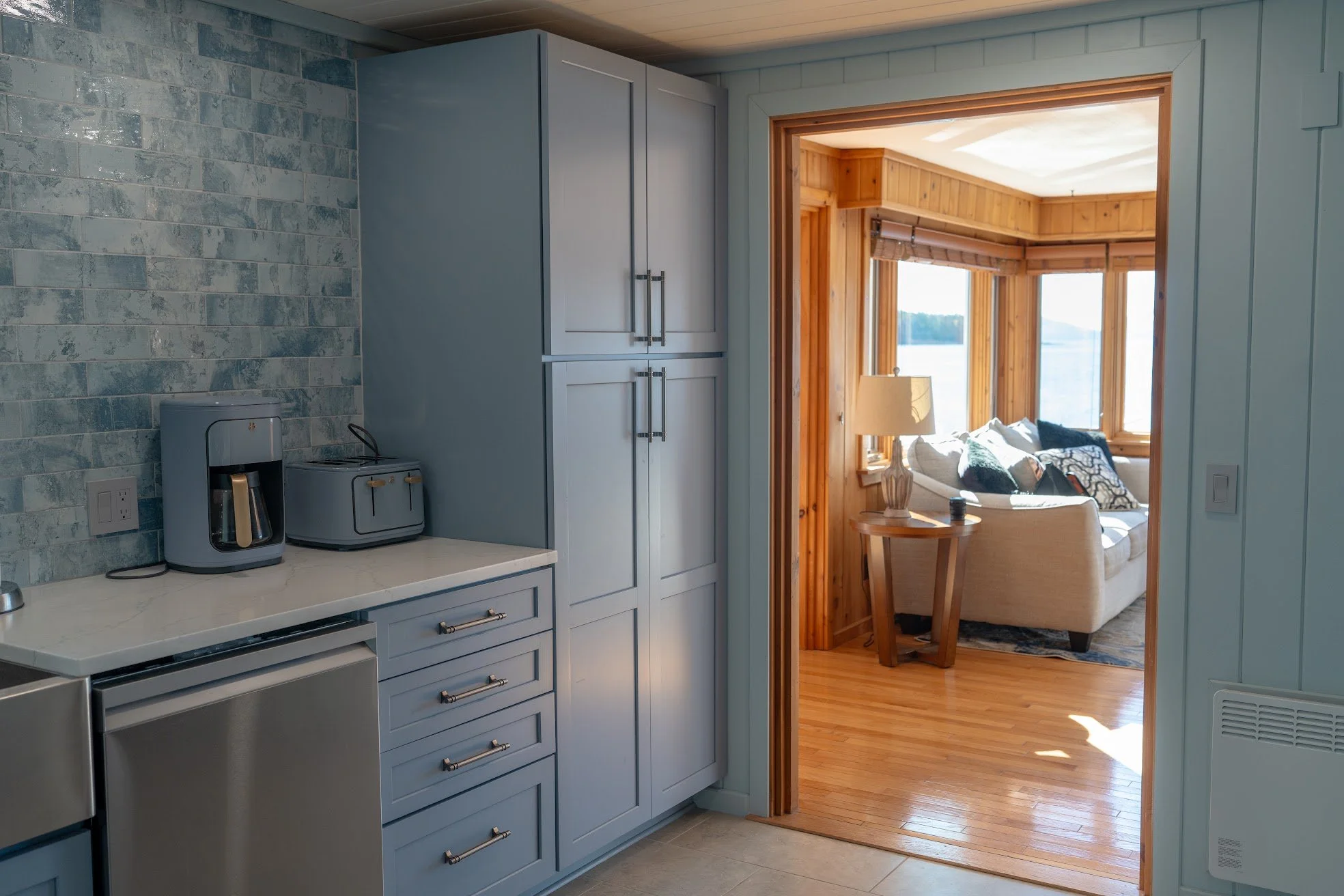 View from a kitchen into a sunlit living room with wooden paneling and large windows overlooking a body of water.