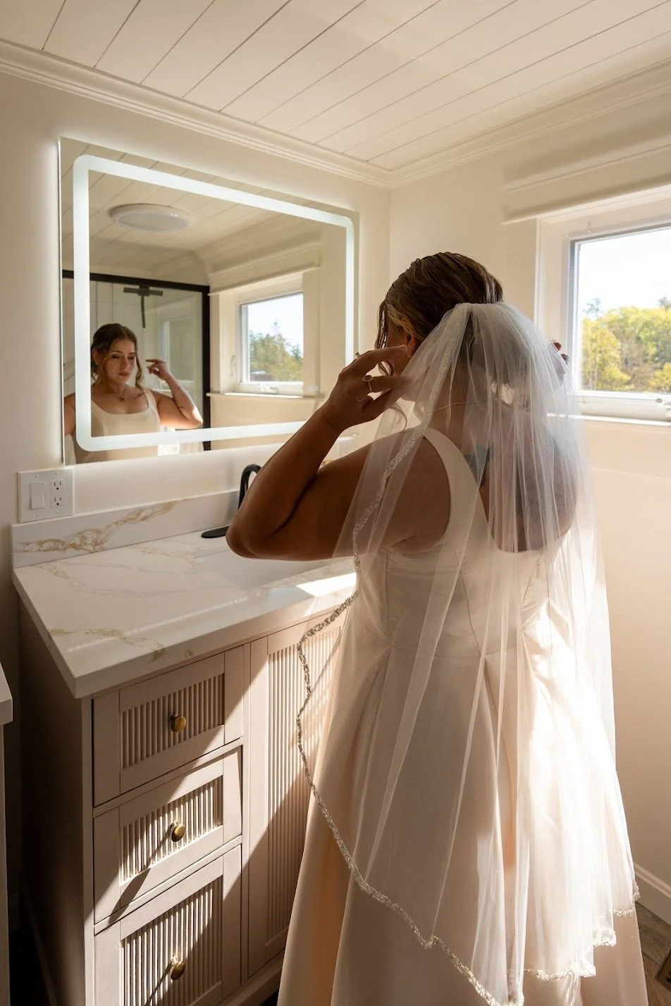 A bride adjusting her veil in front of a mirror in a bright bathroom with a window and a marble countertop.