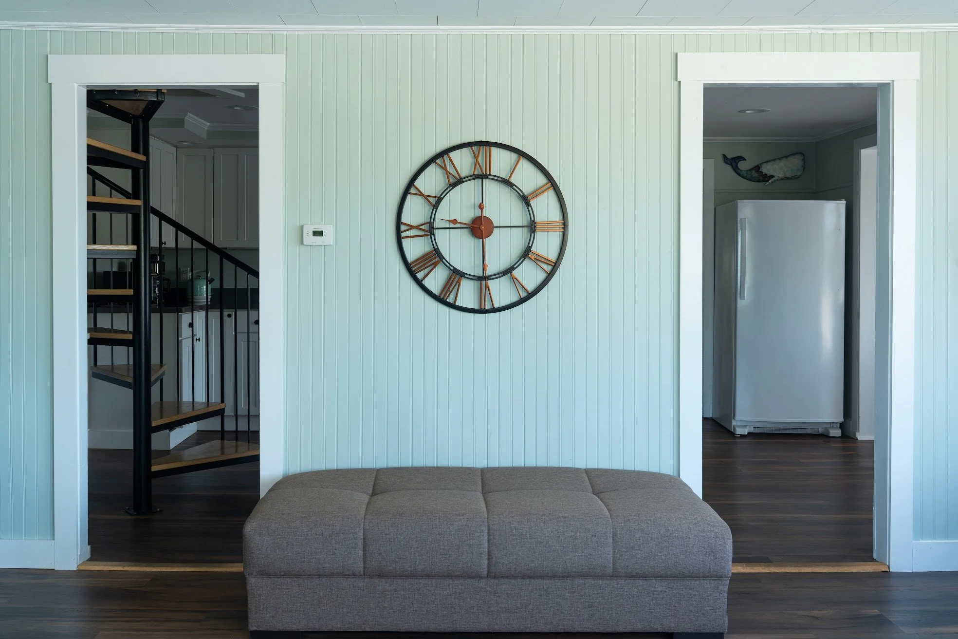 Living room with light green beadboard walls, a large wall clock with Roman numerals, a brown upholstered ottoman, and view into a kitchen with white cabinets and black spiral staircase.