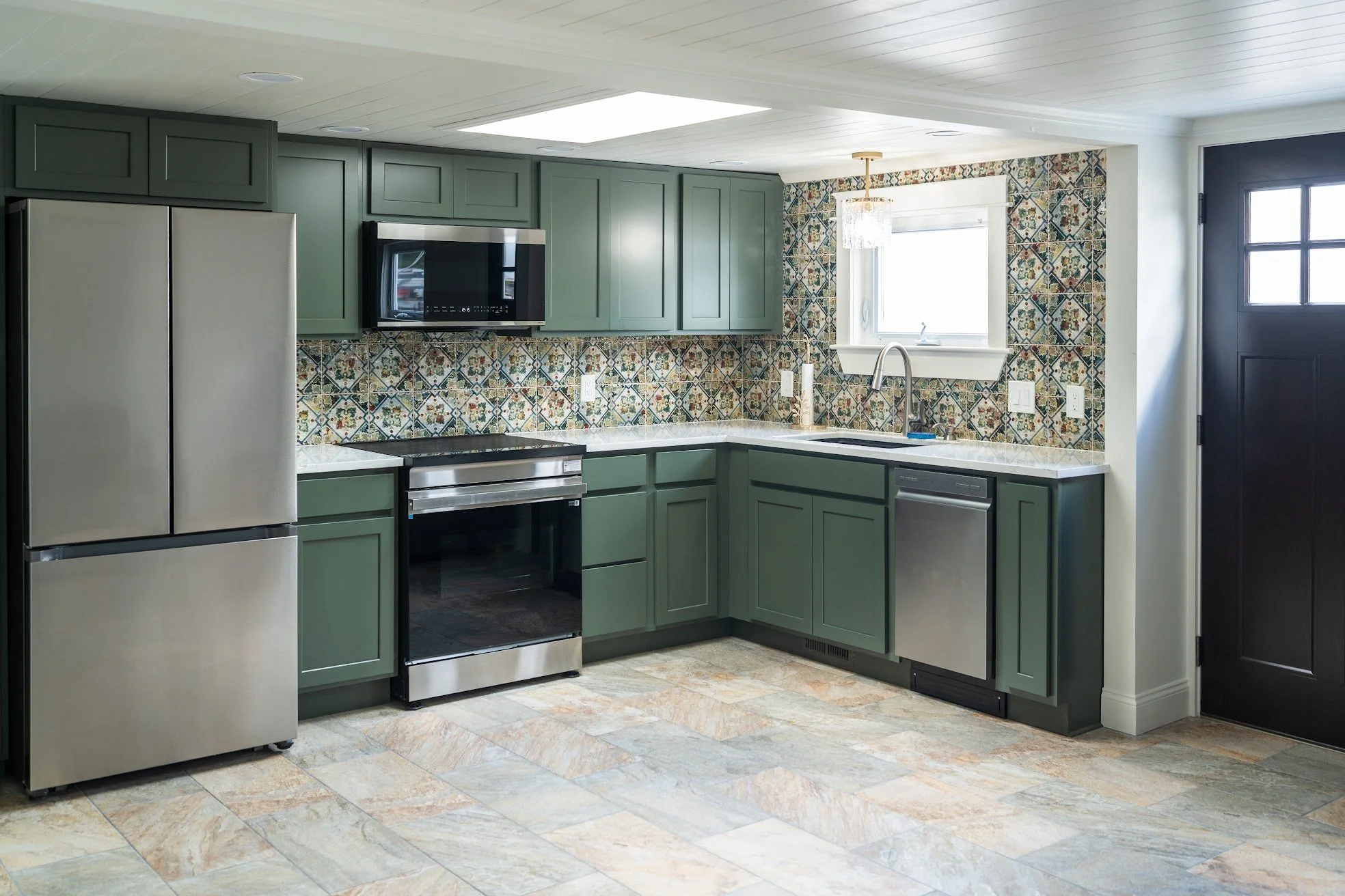 Modern kitchen with green cabinets, patterned tile backsplash, stainless steel appliances, a window above the sink, and a black door.