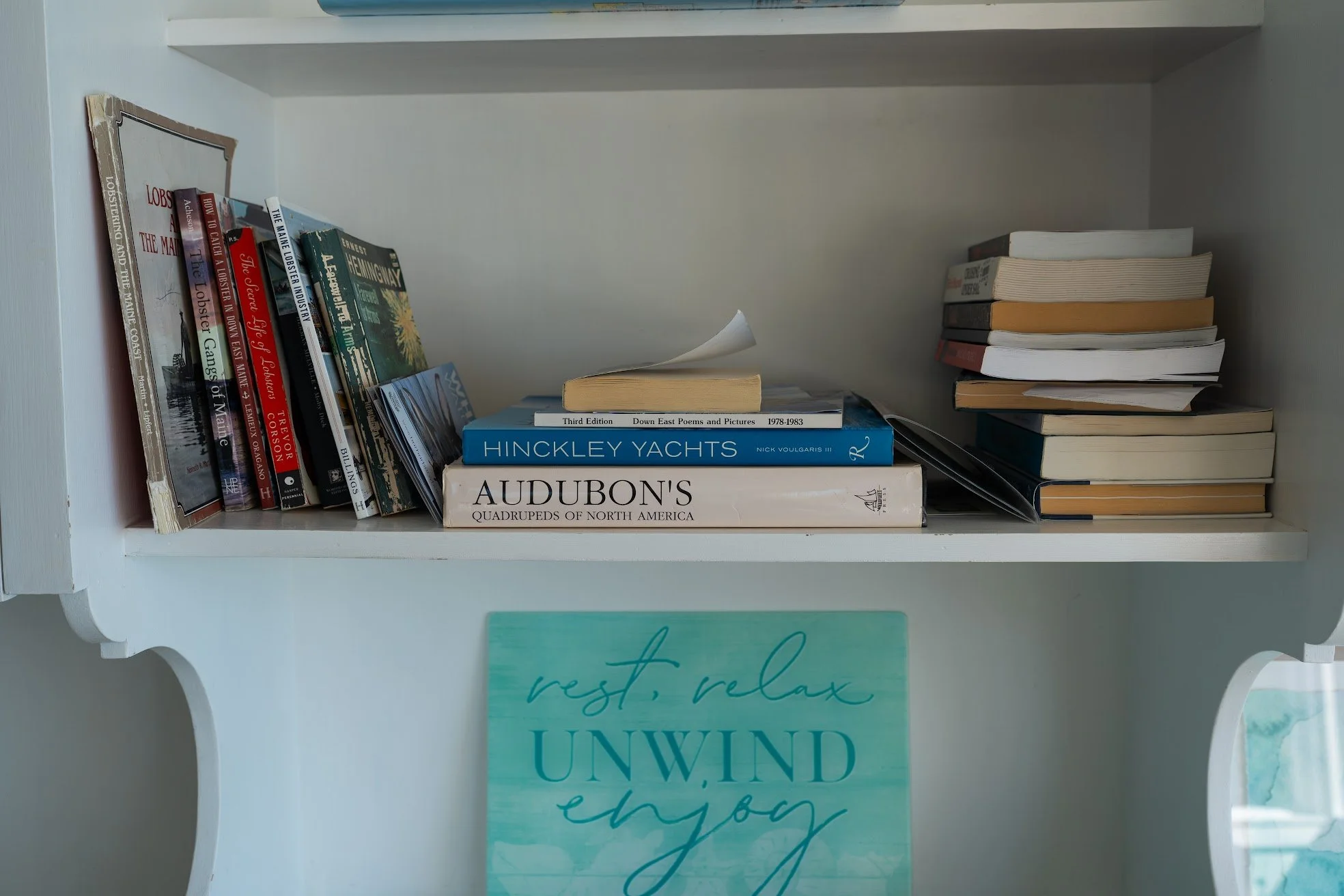 A white bookshelf with books, including titles on yachting, poetry, and North American history, and a decorative sign that reads "rest, relax, unwind, enjoy".