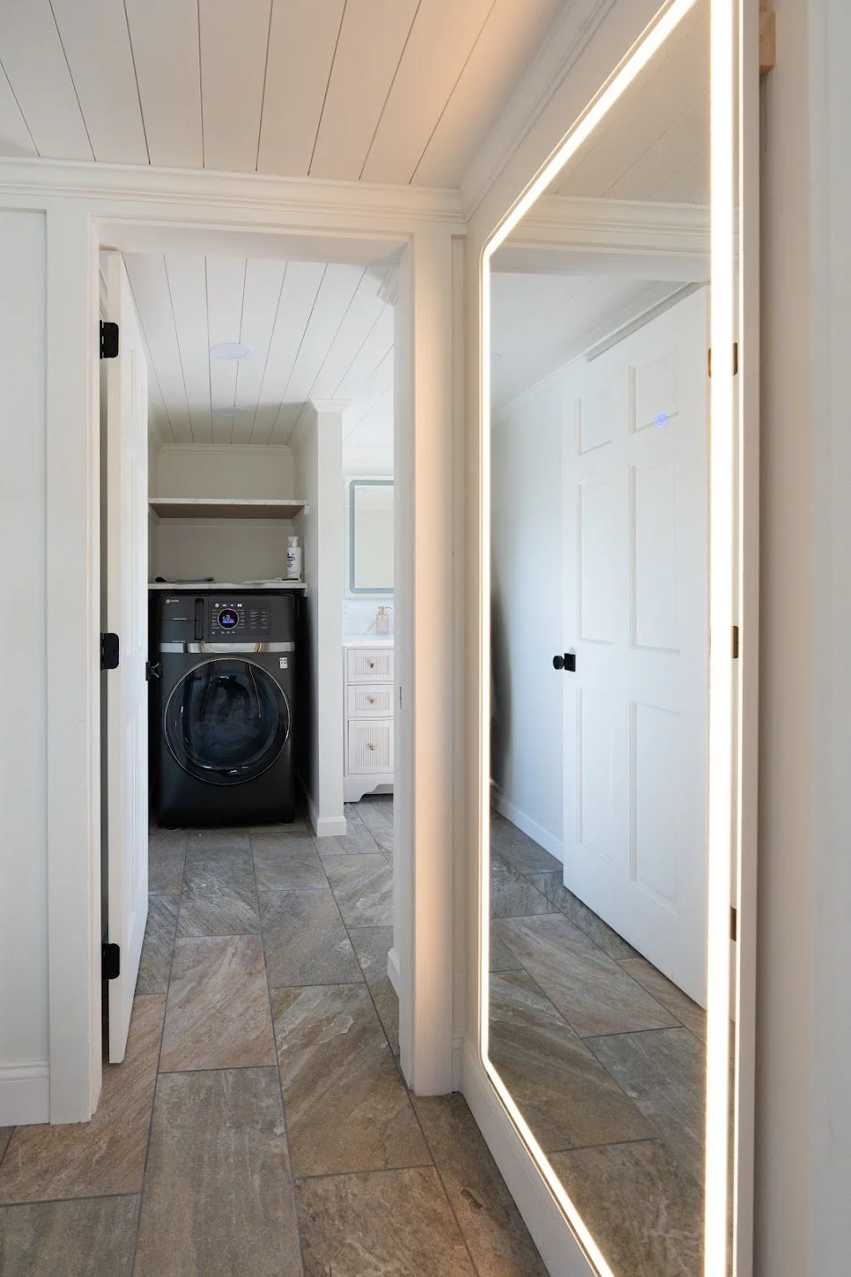 A laundry room with a black front-loading washing machine, white cabinets, a large mirror with an LED light border, and beige tiled floors.