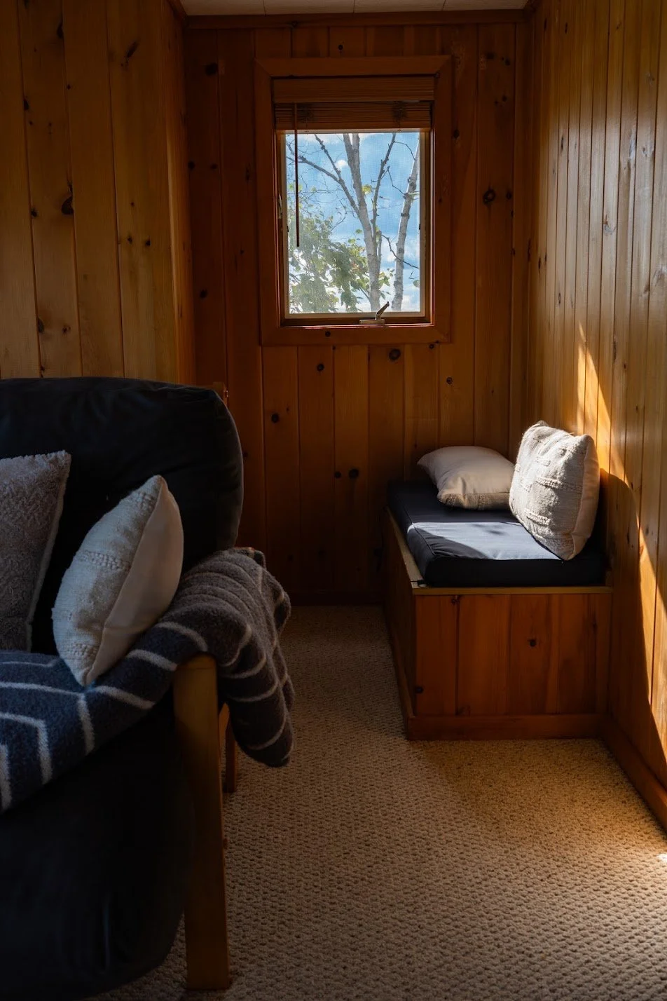 A small cozy room with wood-paneled walls, a window showing a tree outside, a dark couch with pillows and a blanket on the left, and a wooden bench with cushions on the right.