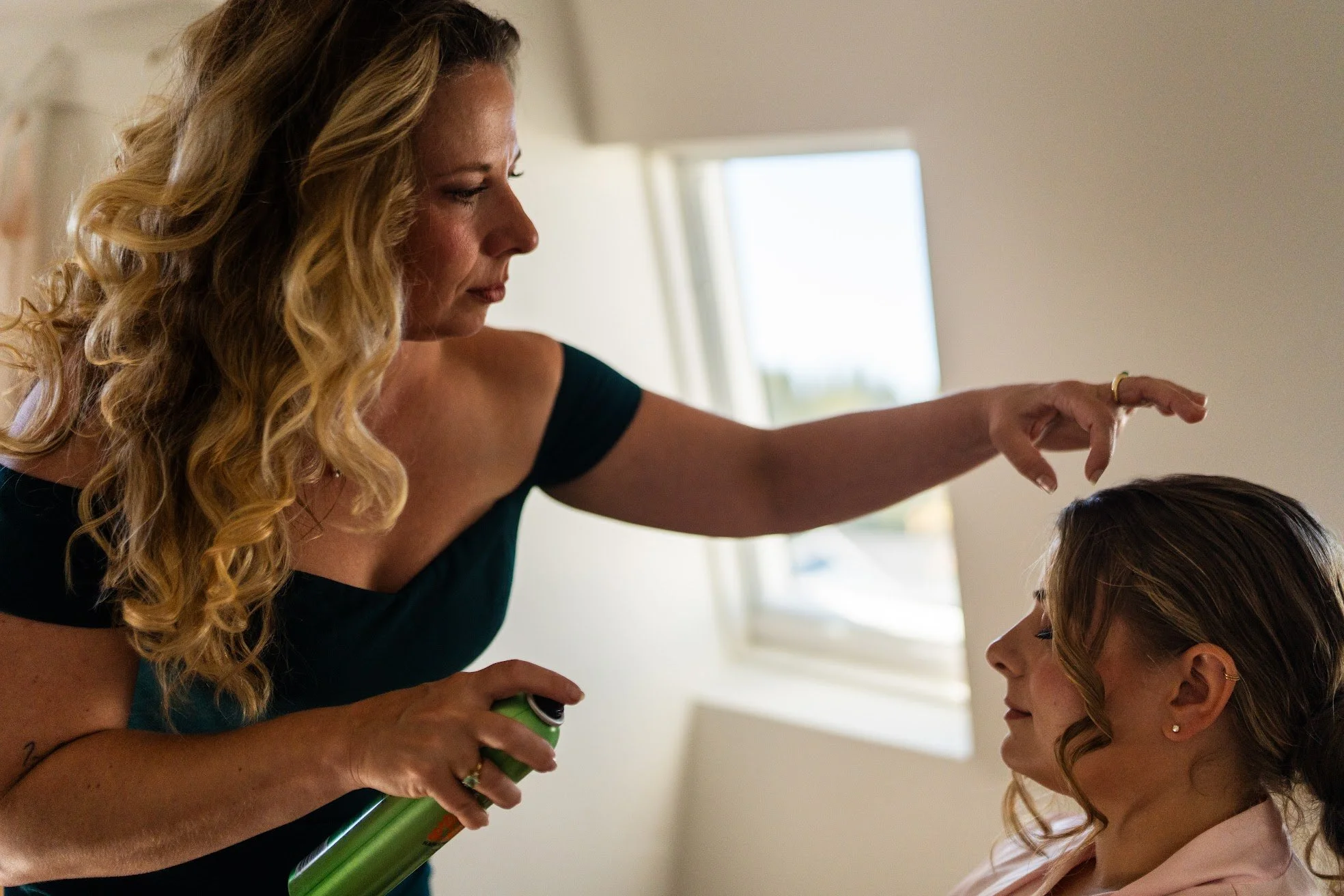 A woman with long blonde hair styling another woman’s hair in a well-lit room.