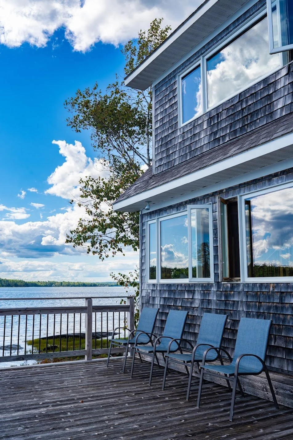 Deck with four blue chairs overlooking a lake, with a gray two-story house with large windows and gray shingled siding, under a partly cloudy sky.
