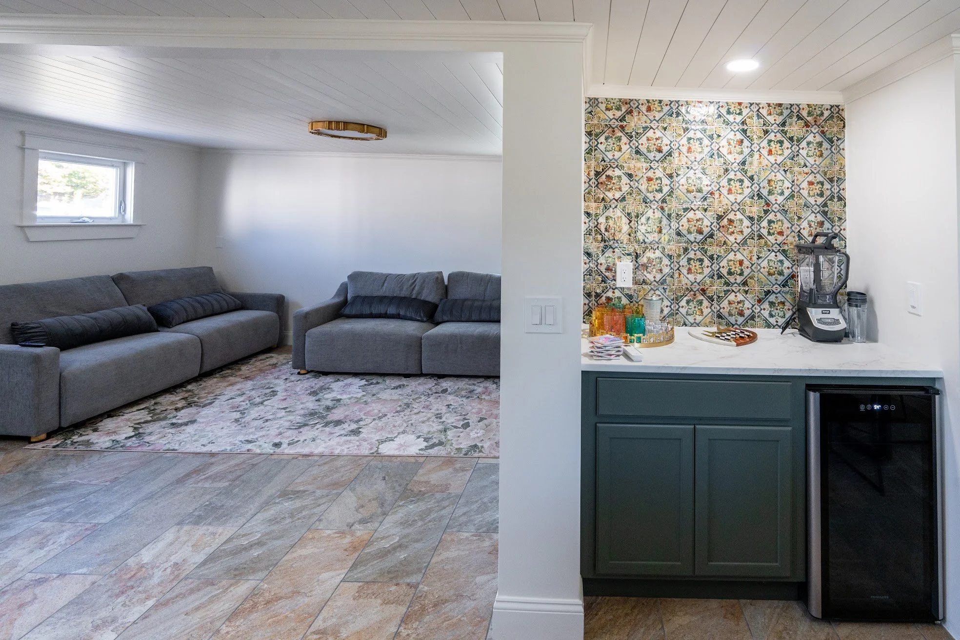Living room with gray sofa, small window, patterned area rug, and wall with recessed lighting; adjacent kitchen with colorful patterned tile backsplash, coffee station, and mini fridge.