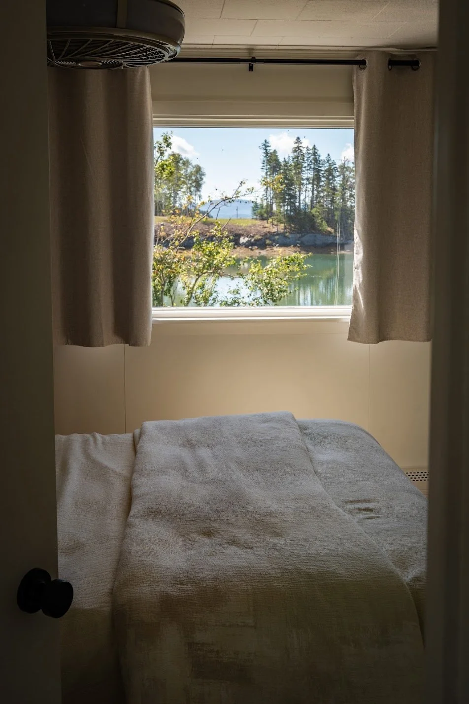 A cozy room with beige curtains, a bed covered with a white textured blanket, and a window showing a scenic view of trees, water, and a clear sky.