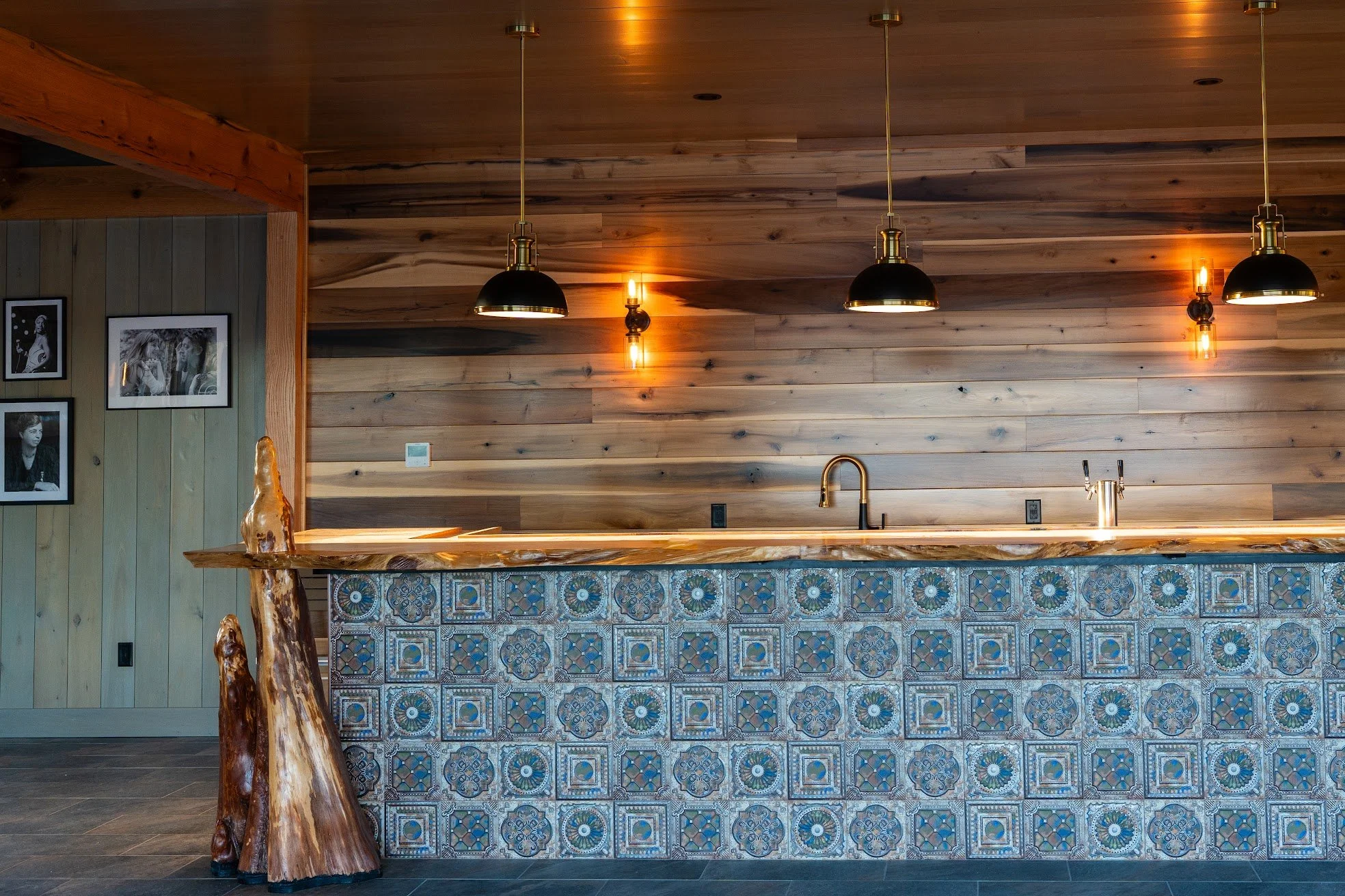 Interior of a modern kitchen with a wooden wall, hanging black and gold pendant lights, a patterned tile front on the counter, and framed black and white photographs on the wall.