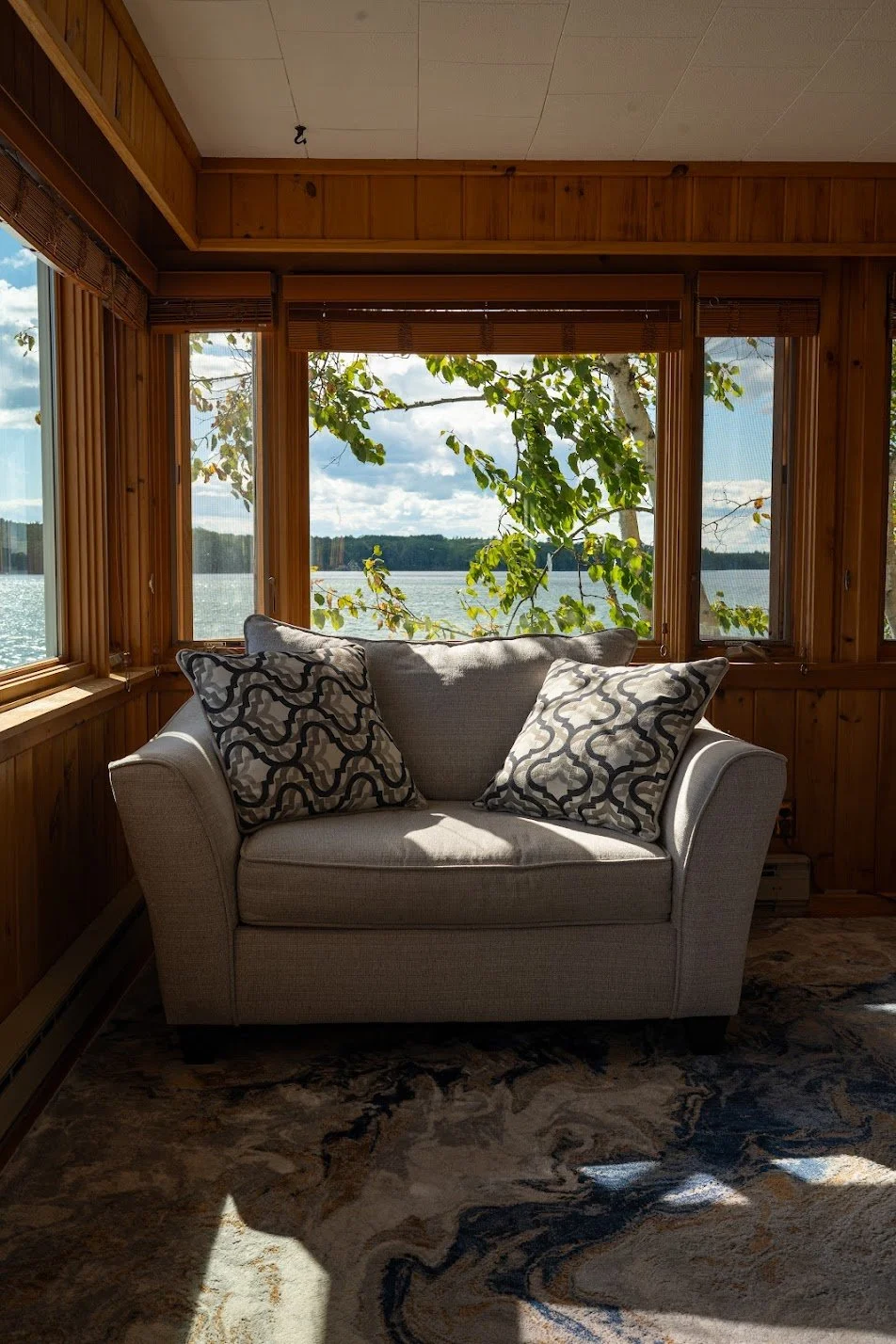 A cozy living room with a beige sofa and two decorative pillows, wooden panel walls, and large windows showing a lake with trees and a partly cloudy sky outside.