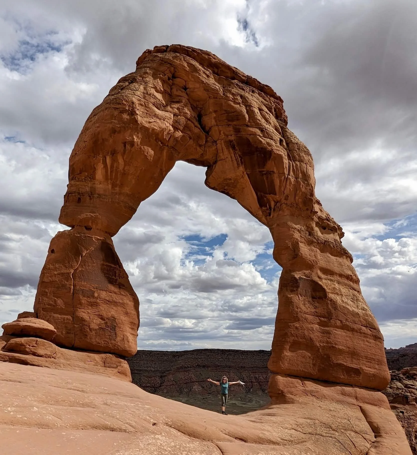 Dr. Maizes standing with arms outstretched beneath a large natural sandstone arch in a desert landscape with cloudy sky.