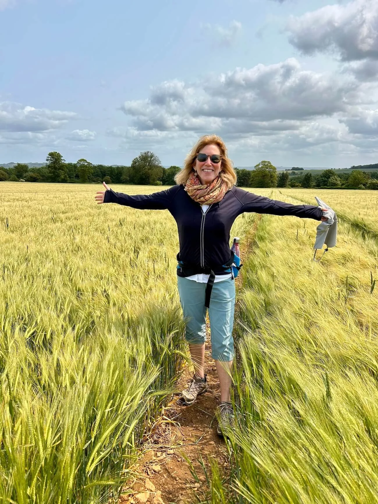 Dr. Victoria Maizes standing in a wheat field with arms outstretched, smiling, wearing sunglasses, a navy jacket, light green pants, and hiking shoes, under a partly cloudy sky.