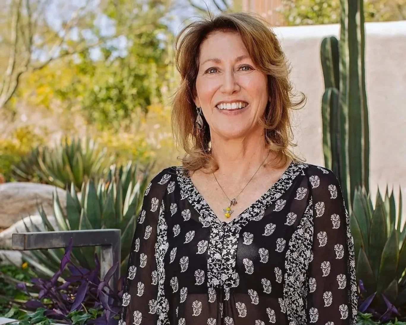 Dr. Victoria Maizes outdoors, wearing a black dress with white patterns, a necklace, and earrings, surrounded by desert plants and trees.