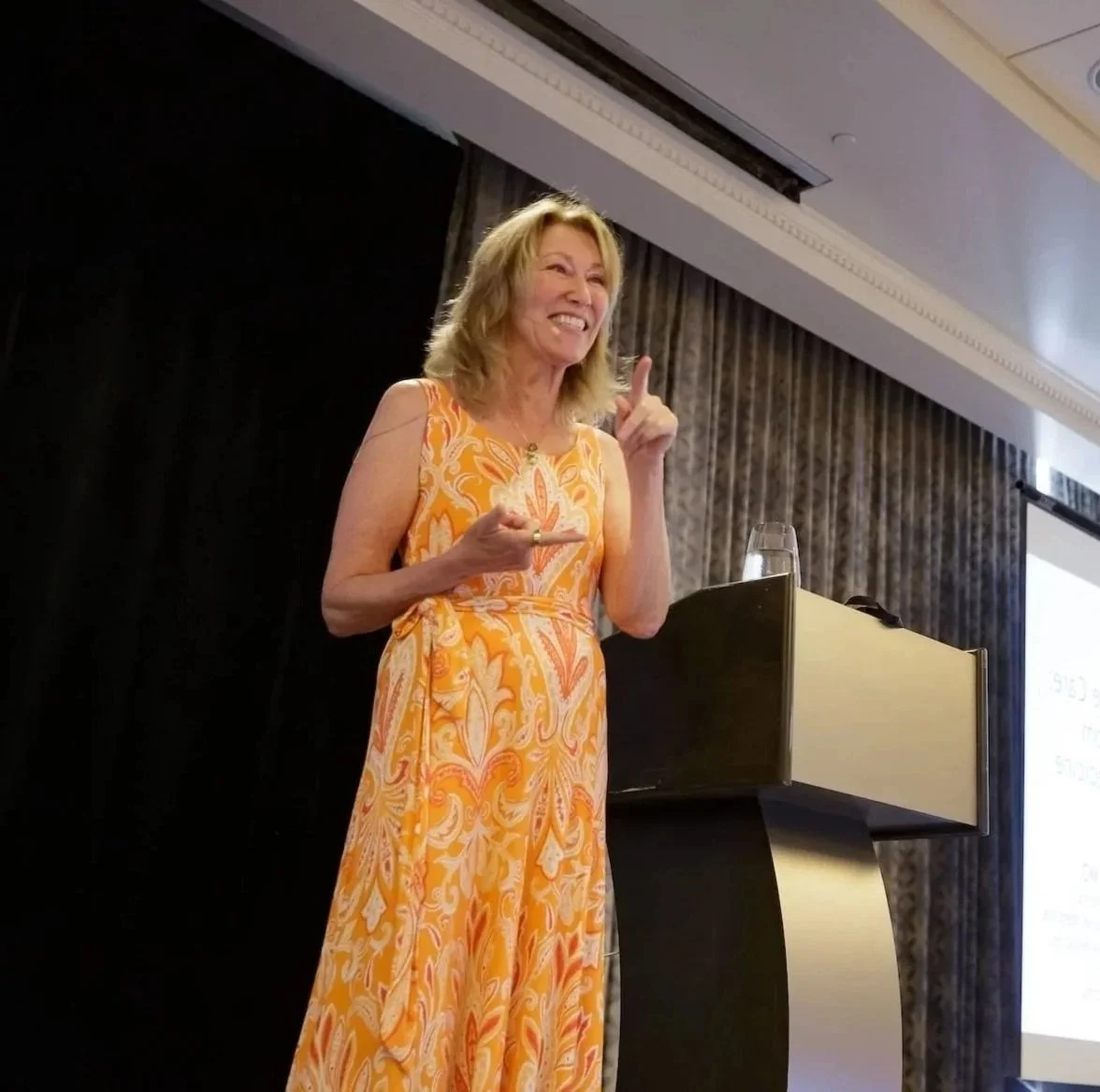 Dr. Maizes teaching in a sleeveless orange and white patterned dress standing at a presentation podium, gesturing with her index finger, beside a glass of water and a screen in the background.