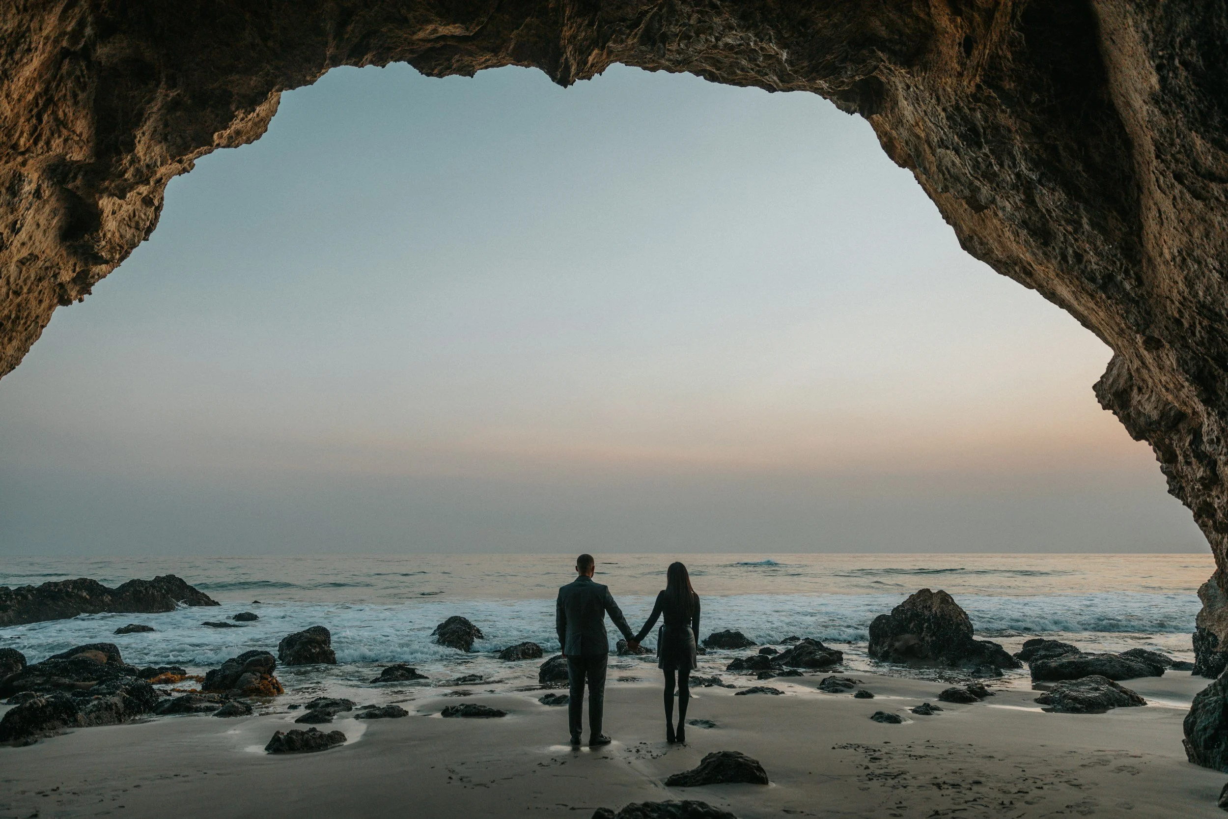Couples holding hands on the beach at dusk under a rock dome