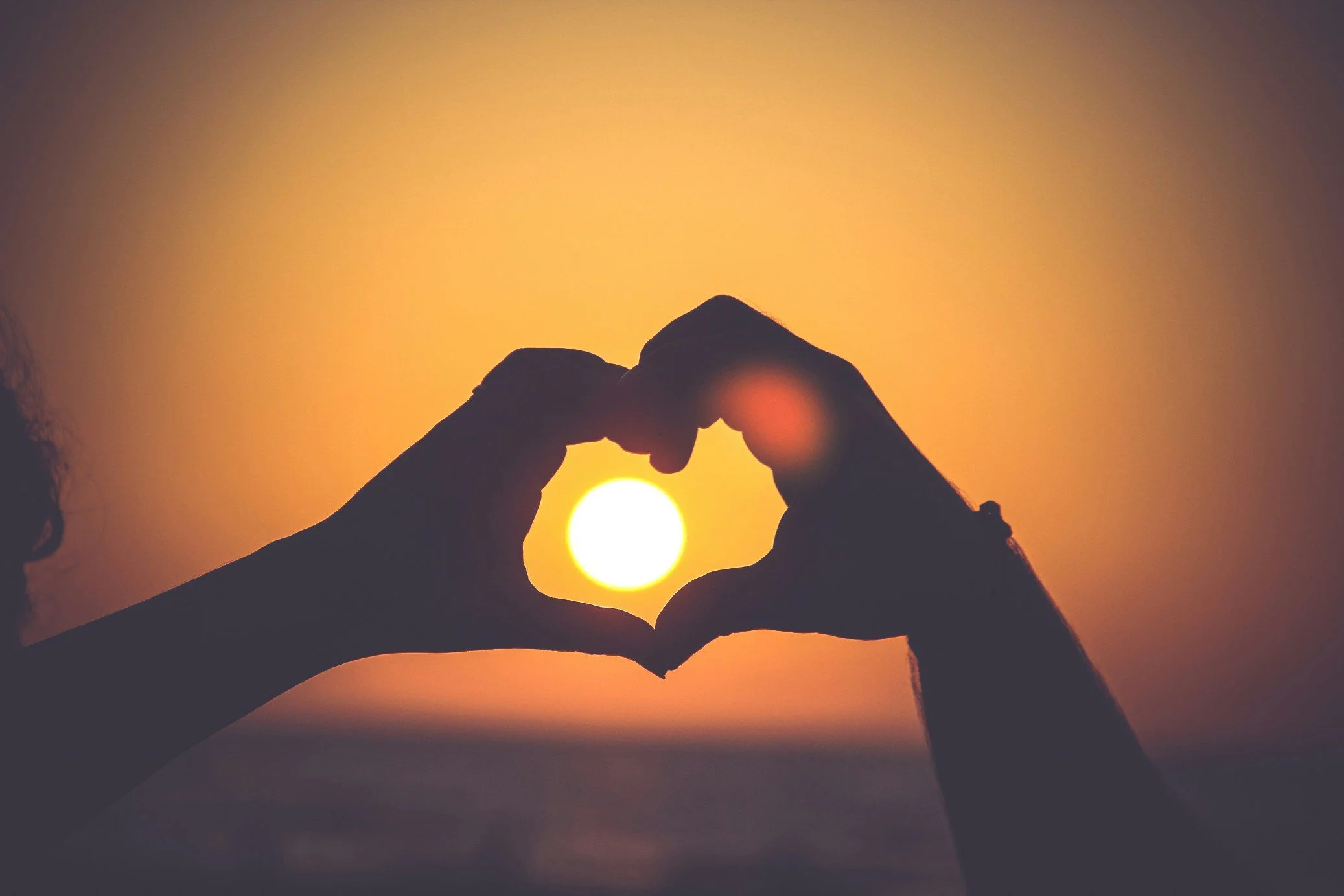 Two hands forming a heart shape around the setting sun at sunset.