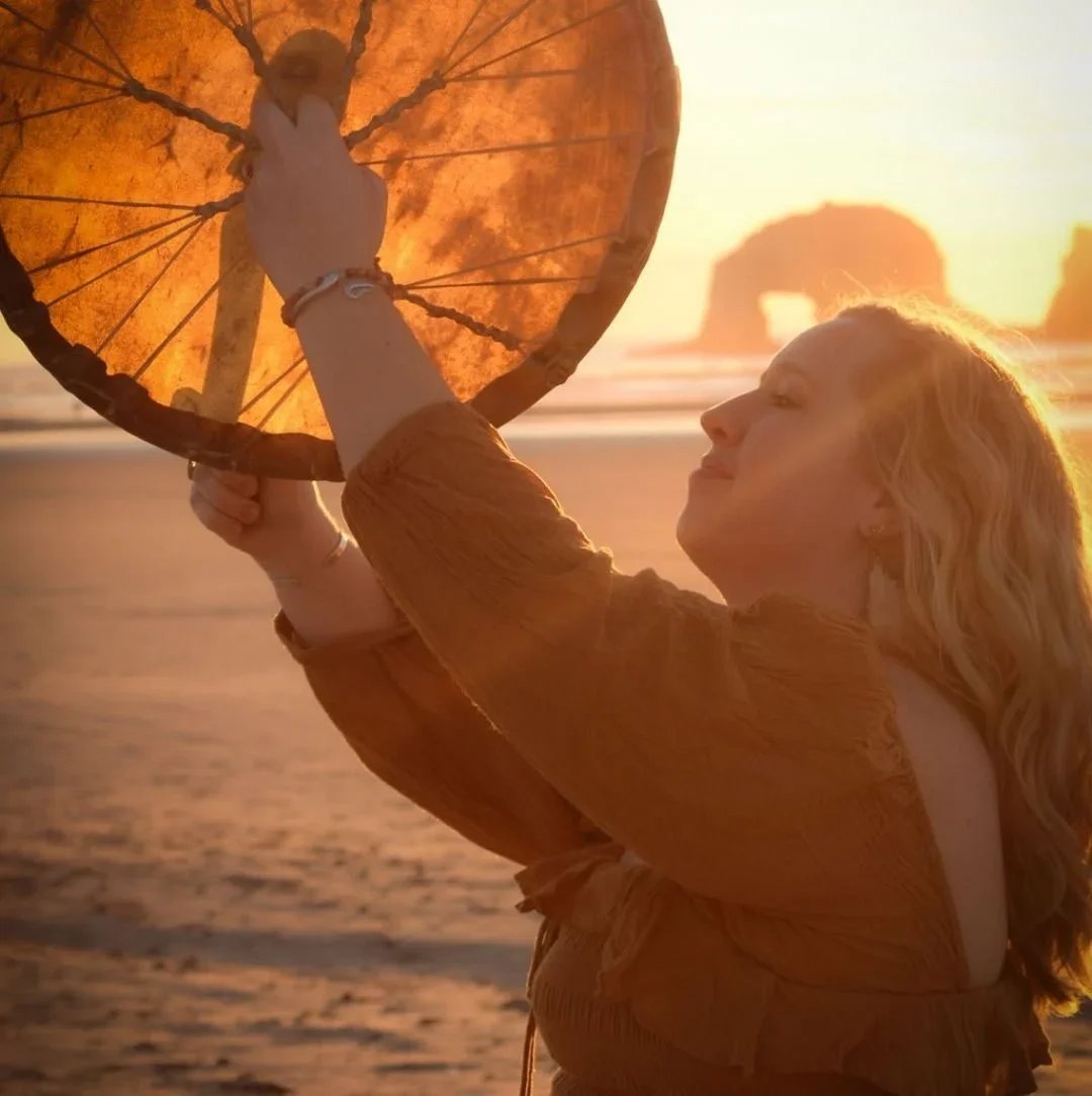 McKinley Merchant, LCSW is holding a large drum up to the sun on a beach during sunset