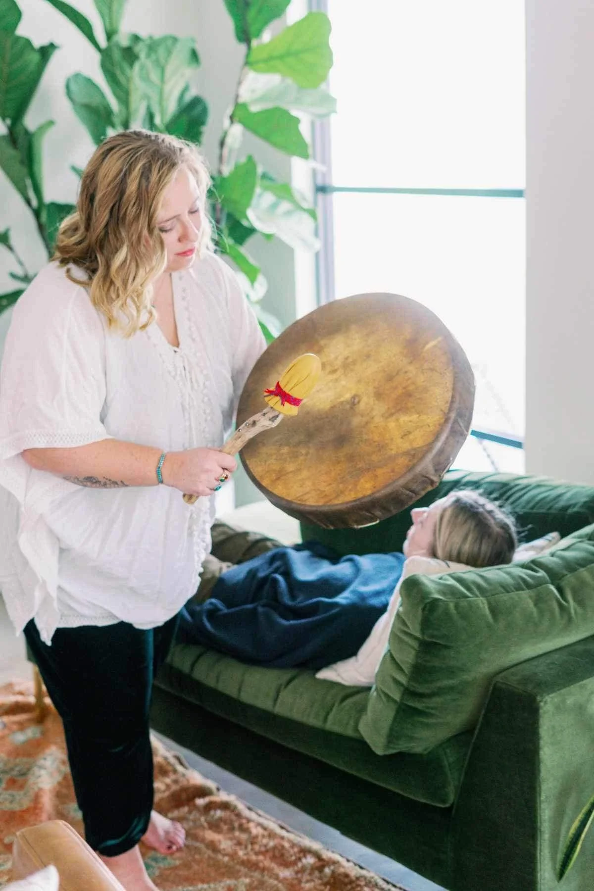 A woman standing next to a person lying on a green velvet sofa, holding a large drum and a drumstick with a decorative end. The woman is wearing a white top, and the person on the sofa is lying down with headphones on, in a bright room with large green plants.