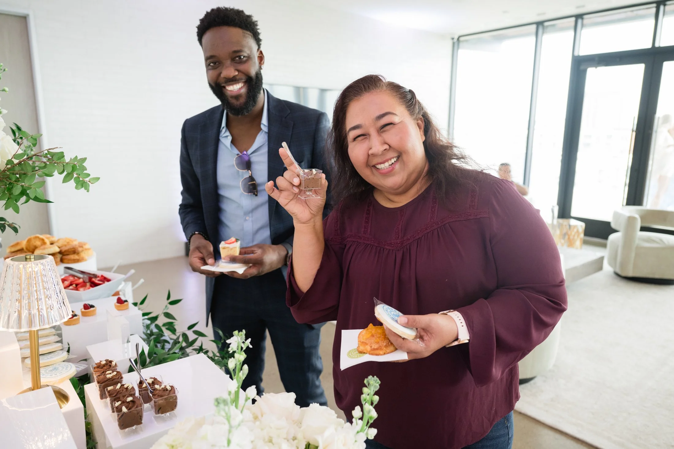 Two people smiling and enjoying desserts at a social gathering or celebration, with a table full of cakes, pastries, and chocolates in a bright, modern room.
