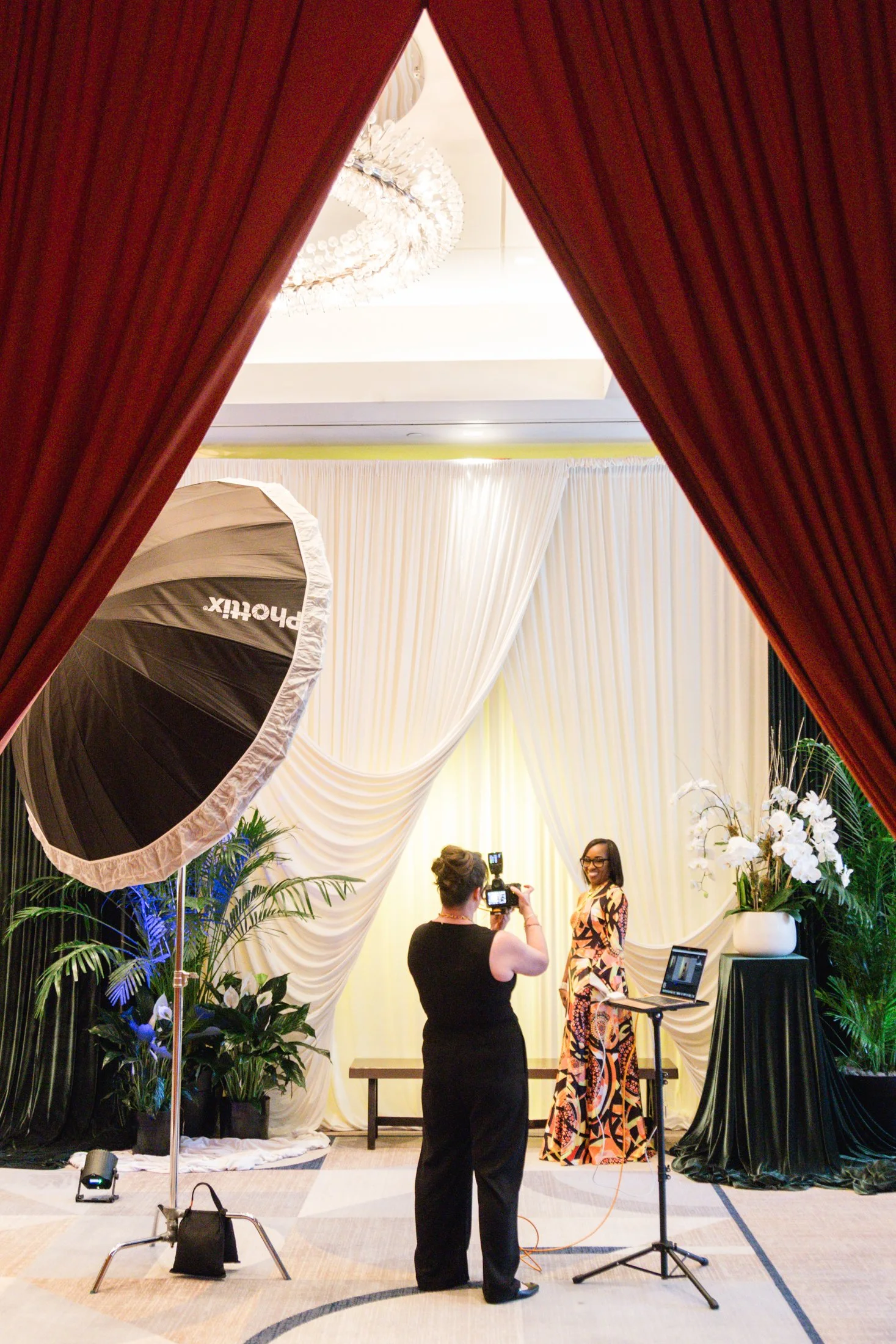 A woman is taking a photo of another woman standing in front of a white curtain backdrop with drapes at an event or photo shoot. There is professional lighting equipment and a laptop on a stand nearby, and the scene includes decorated plants and flow