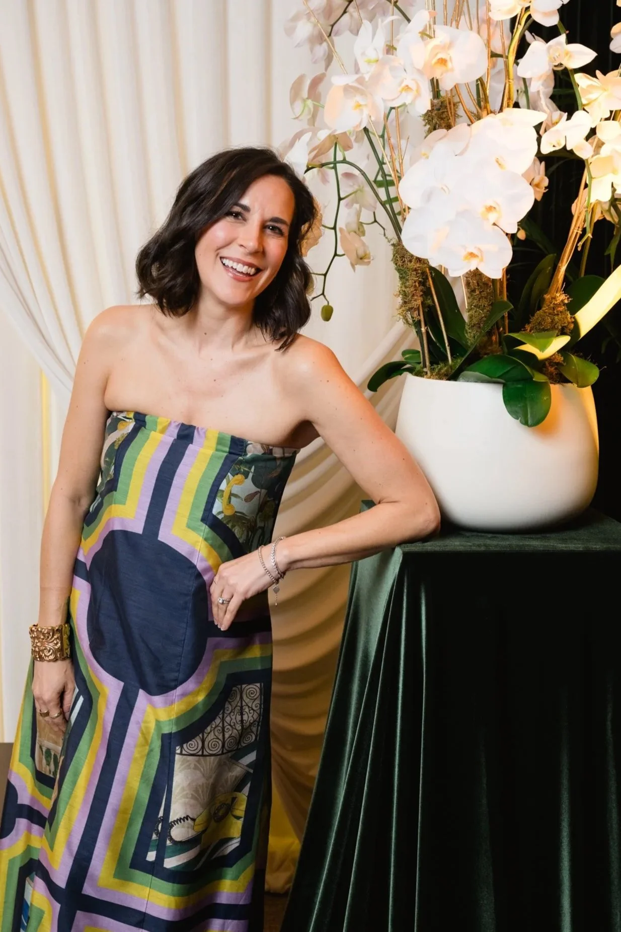Woman in a colorful strapless dress smiling and posing next to a large white vase with white orchids.