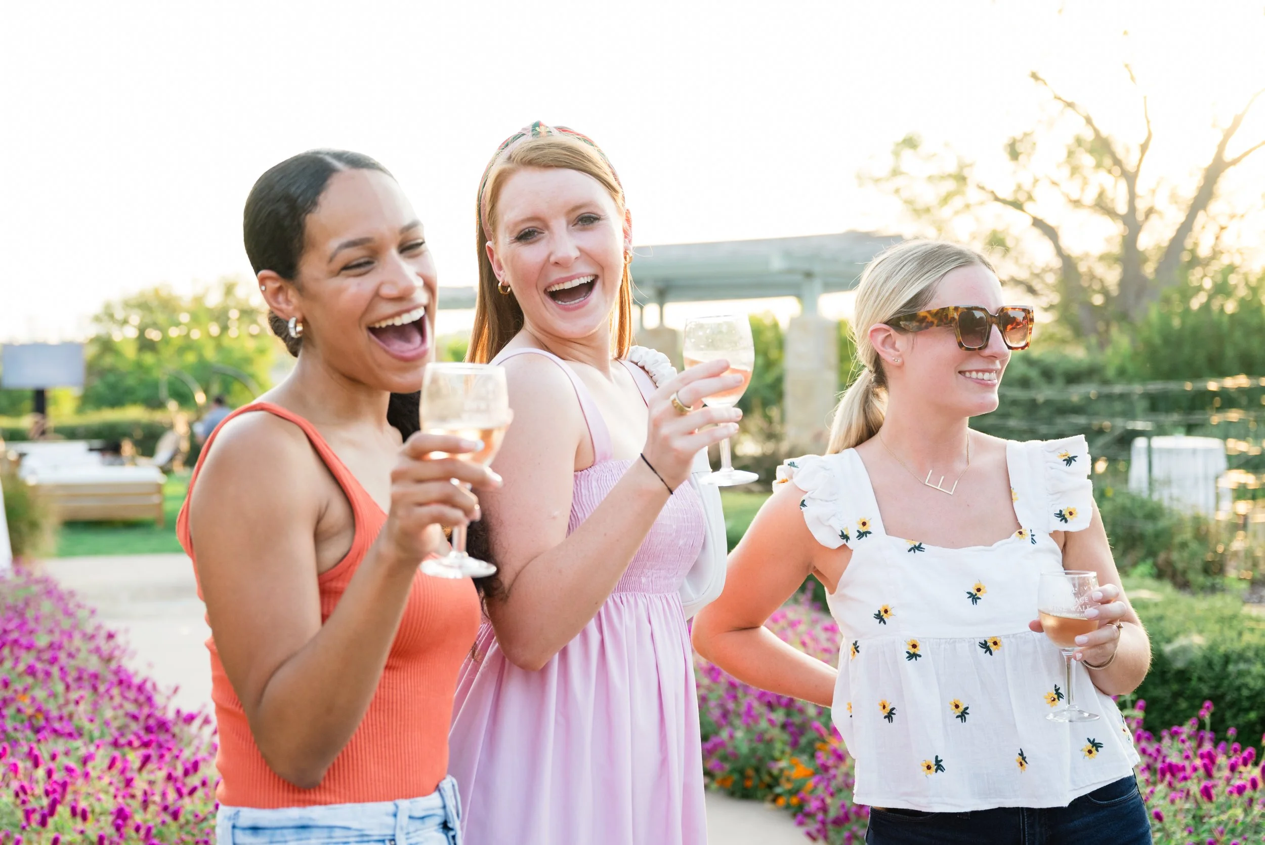 Three women smiling and holding wine glasses outdoors during sunset, surrounded by colorful flowers and greenery.
