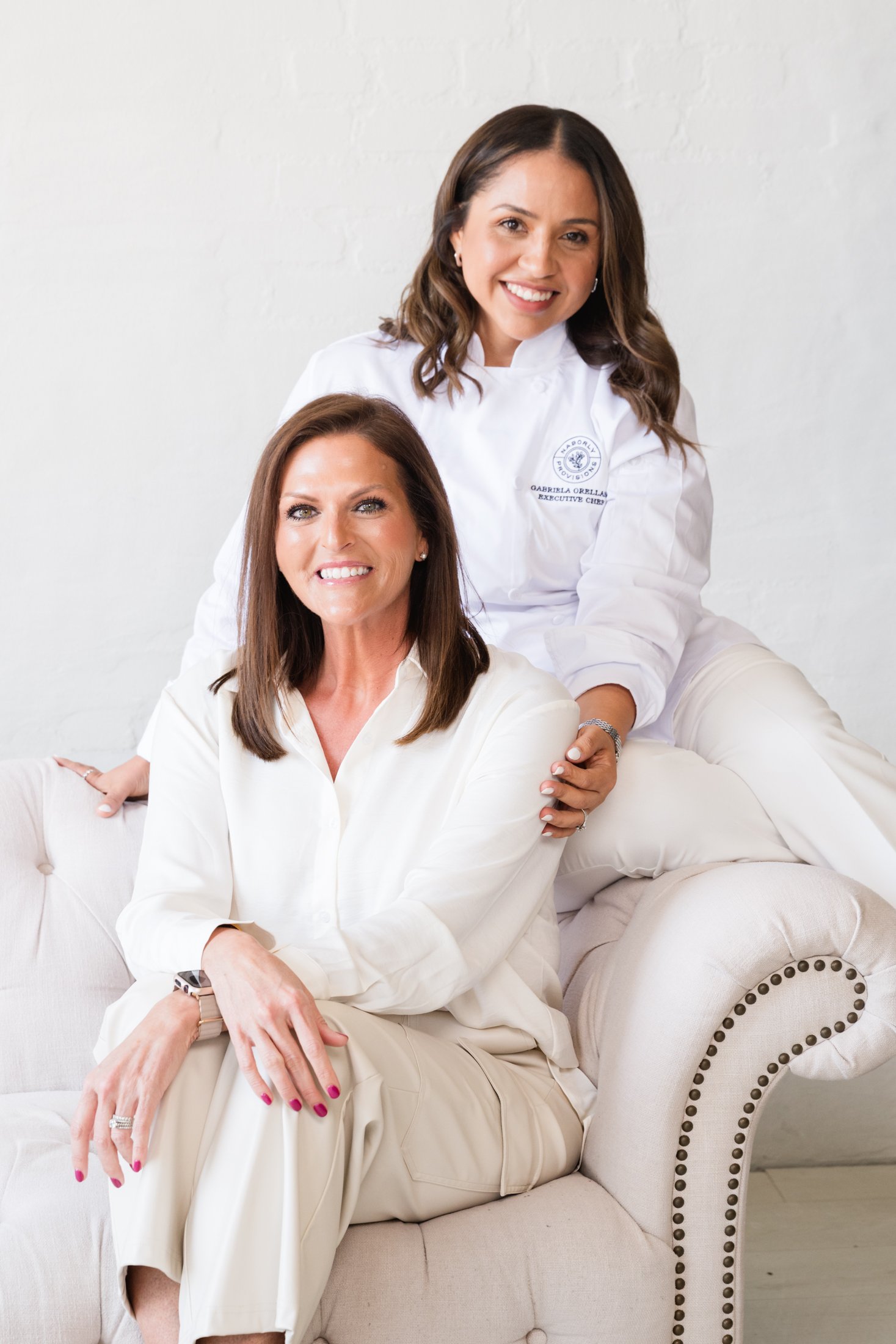 Two women, one seated on a cream-colored sofa and the other sitting on the back of it, smiling in a bright, white room. The woman on the sofa has long brown hair, wearing a white blouse, with rings and a watch on her left hand. The woman behind has s