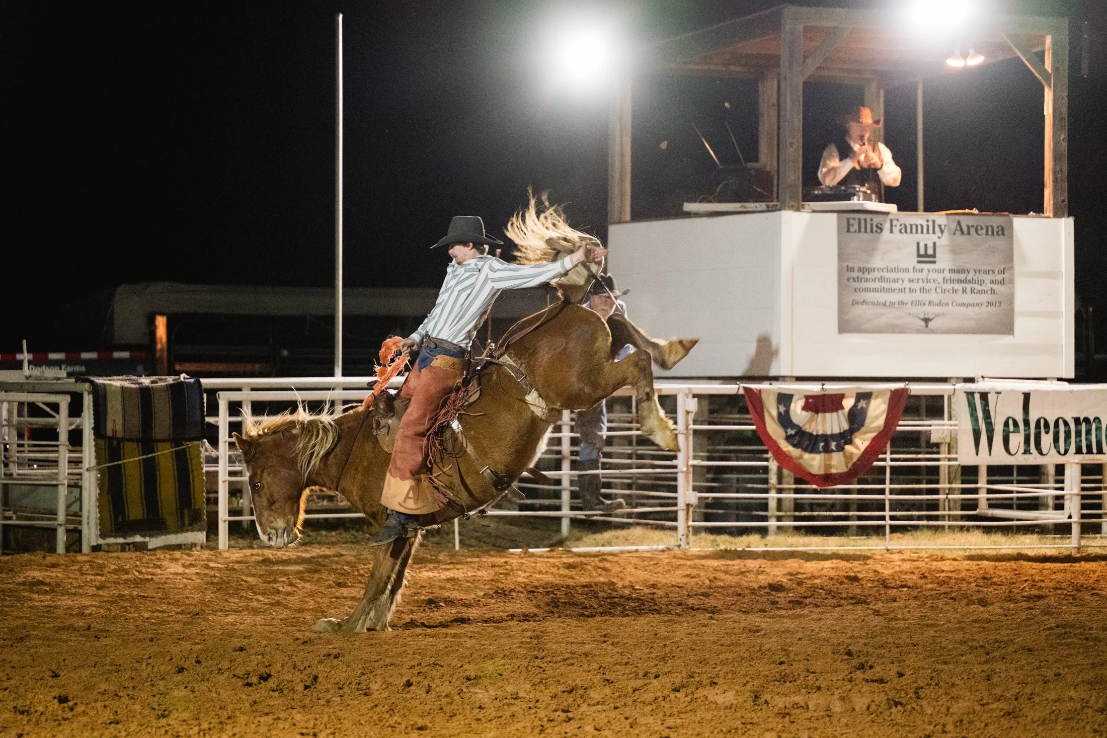 A cowboy riding a bucking horse in a rodeo arena at night, with a person in the background on a platform with a DJ.