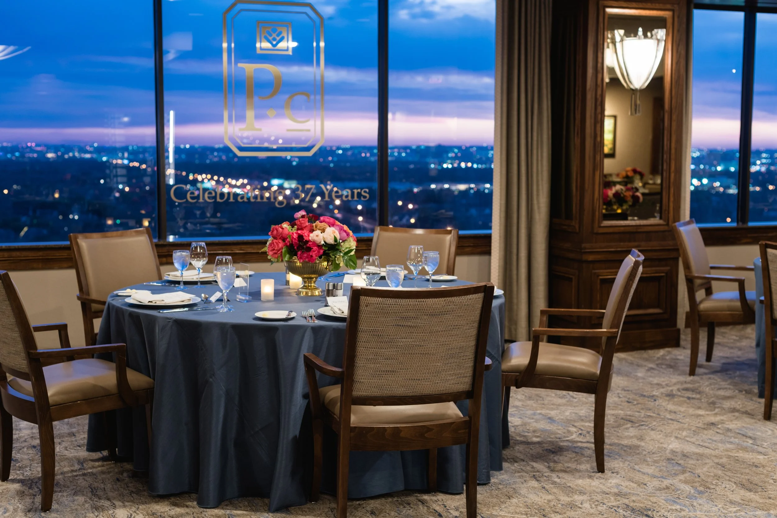 A round table with a dark blue tablecloth, pink and white flower centerpiece, and table settings, in a high-rise restaurant with large windows showing city lights and a sunset sky, celebrating 37 years.
