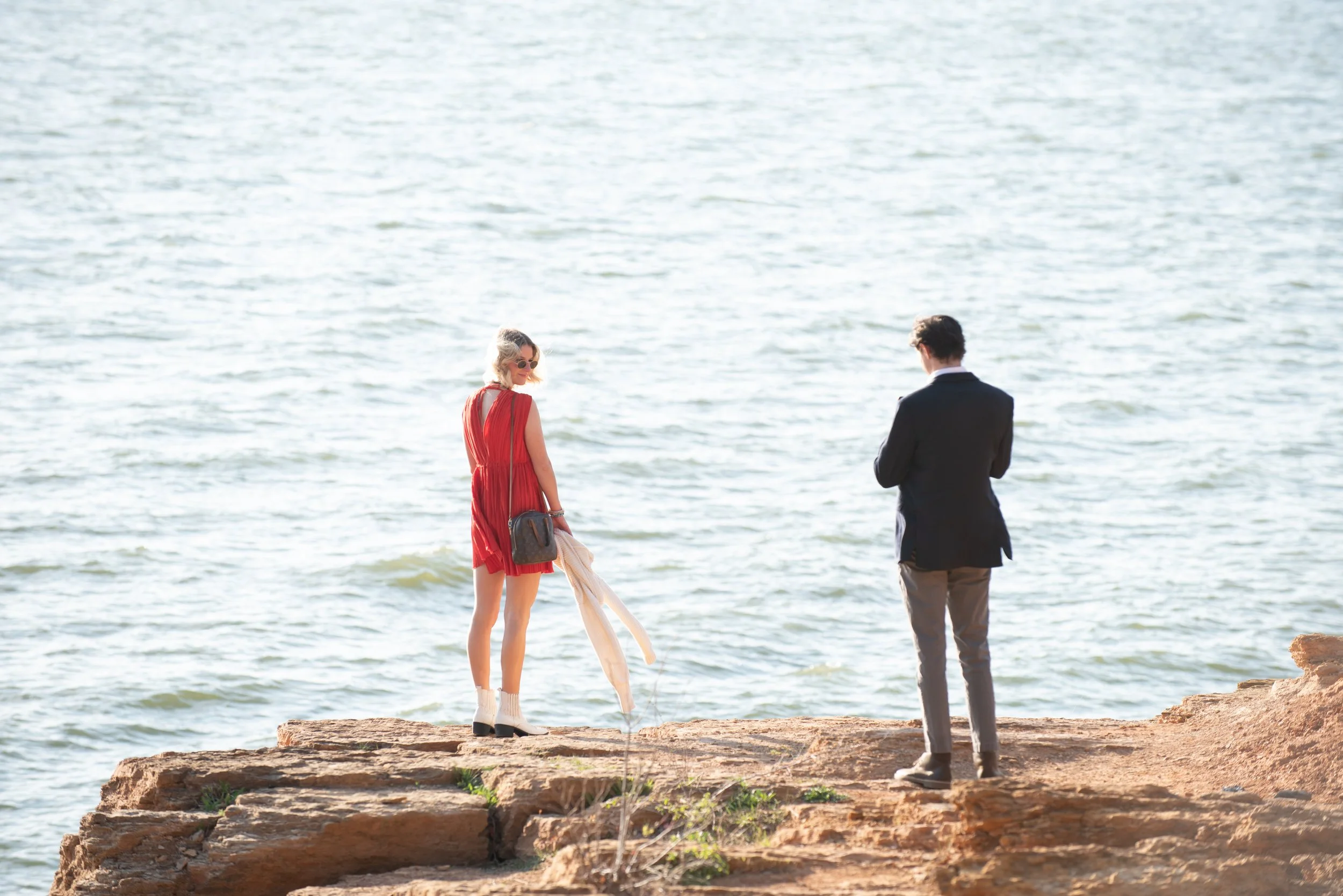 A woman in a red dress with sunglasses and a man in a suit with gray pants are standing on a rocky shoreline by the water, facing each other.