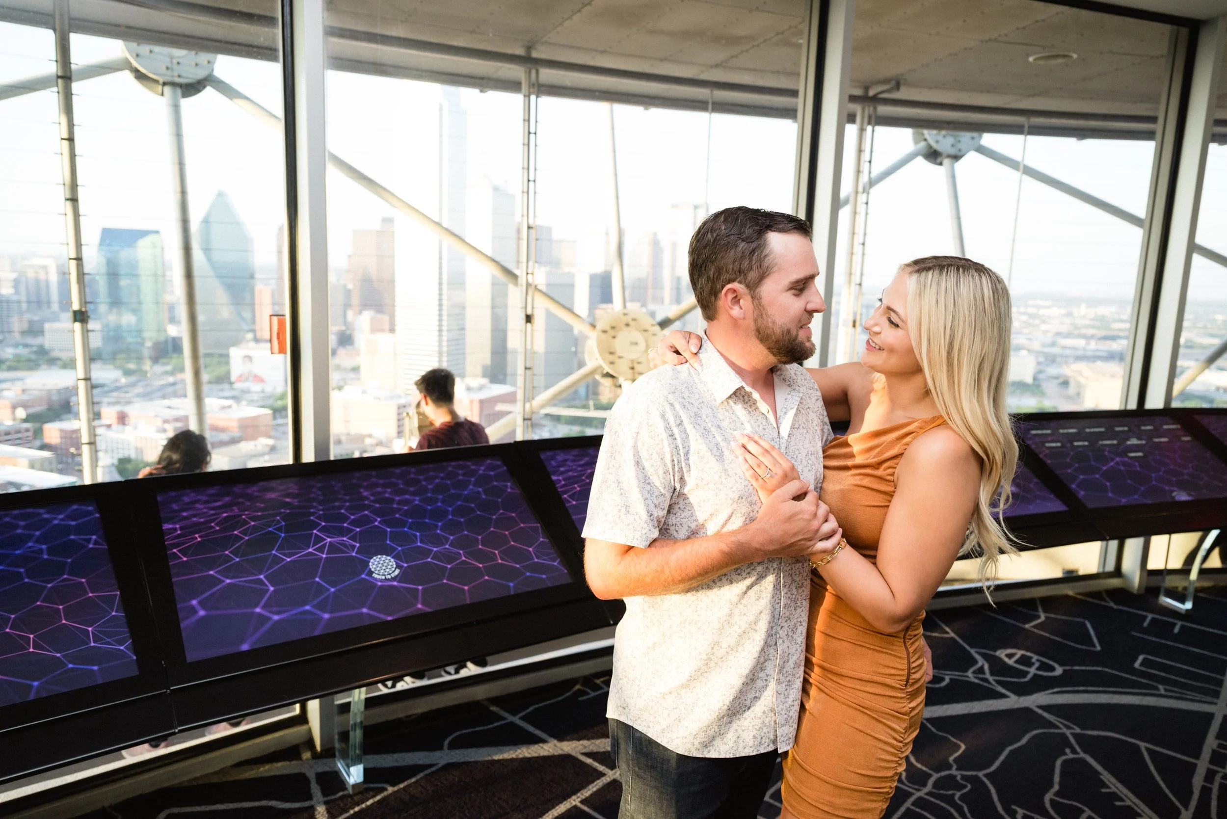 A couple dancing and smiling on an observation deck with large windows and view of the city skyline.