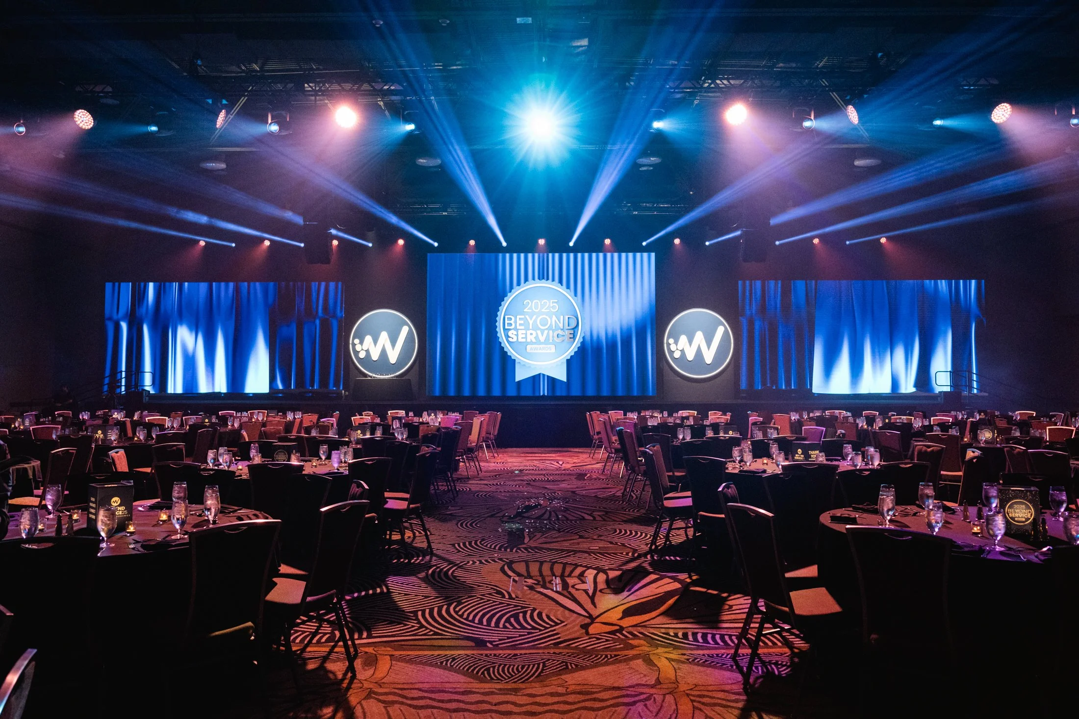 Empty banquet hall with a stage, large screens displaying the '2025 Beyond Service Awards' logo, and round tables set with glasses and silverware, illuminated by blue and purple stage lights.