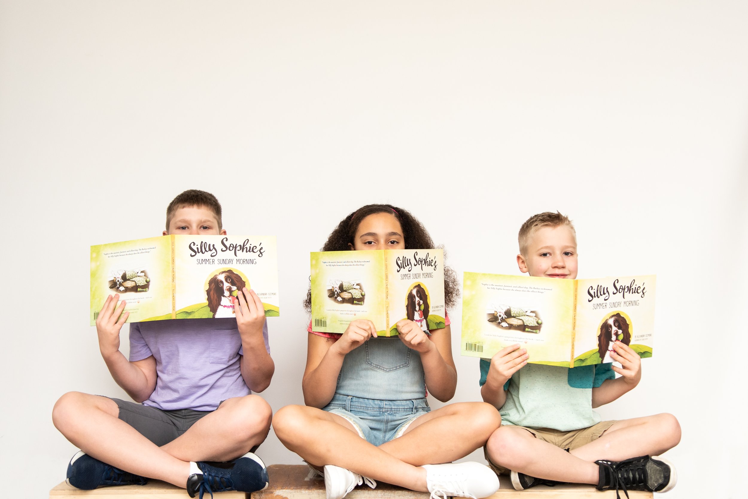 Three children sitting cross-legged on a wooden bench, each holding a children's book titled 'Silly Sophie's Summer Sunday Morning' in front of their faces, against a plain white wall.