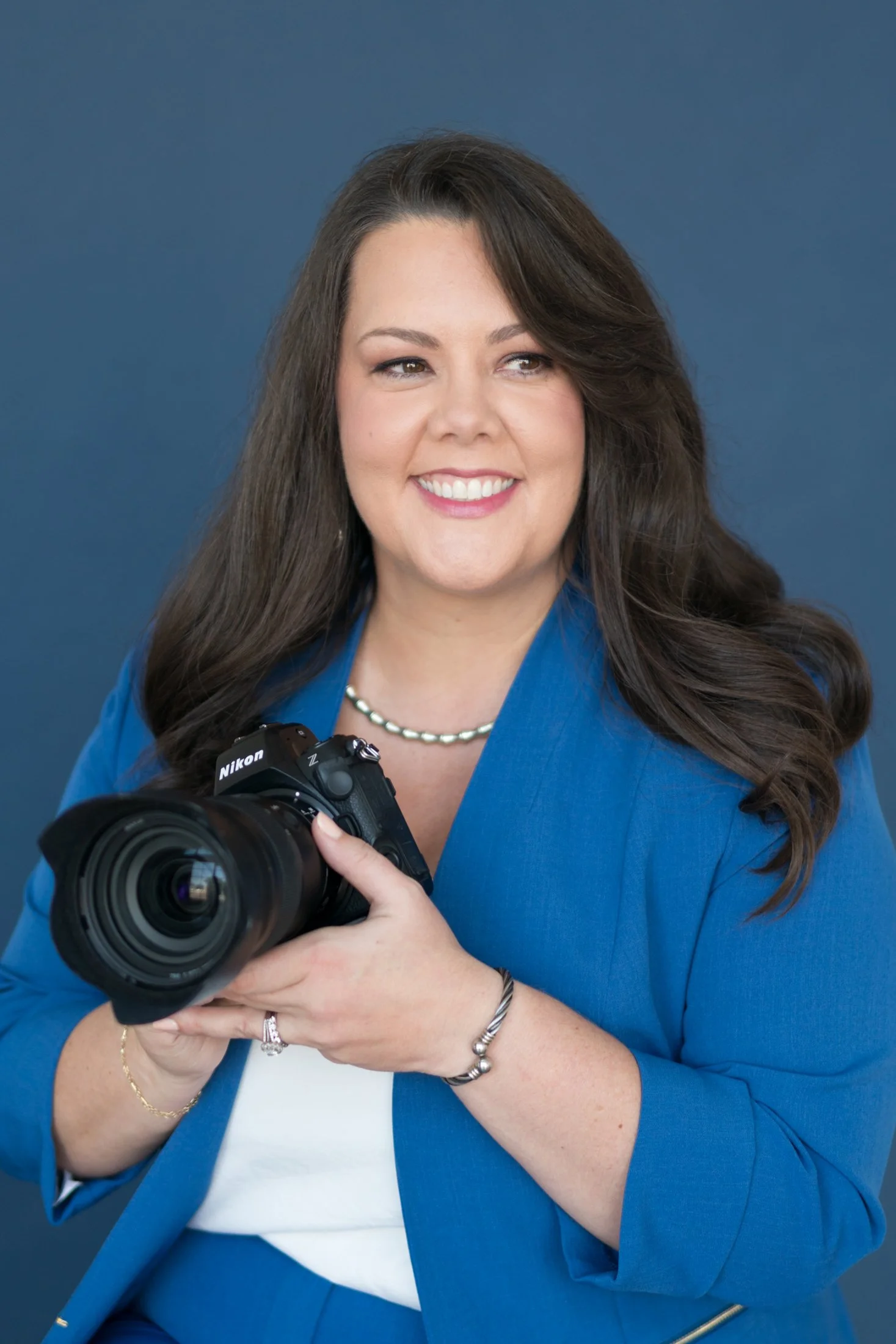A smiling woman with long brown hair wearing a blue blazer, holding a Nikon camera, standing against a solid dark blue background.