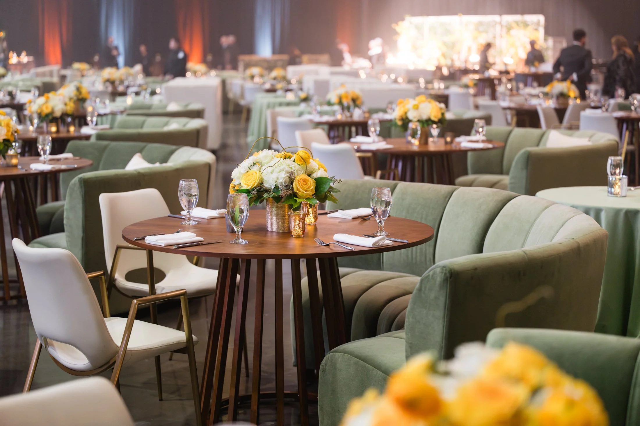 Elegant event space with round tables decorated with floral centerpieces, surrounded by green and white cushioned seating and white chairs, ready for a formal gathering.