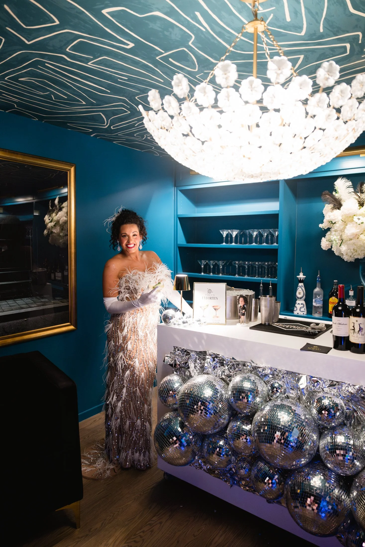 Woman in a glamorous feather and beaded gown standing at a bar decorated with disco balls and silver foil, under a large floral chandelier, in a room with blue walls and a mirror.