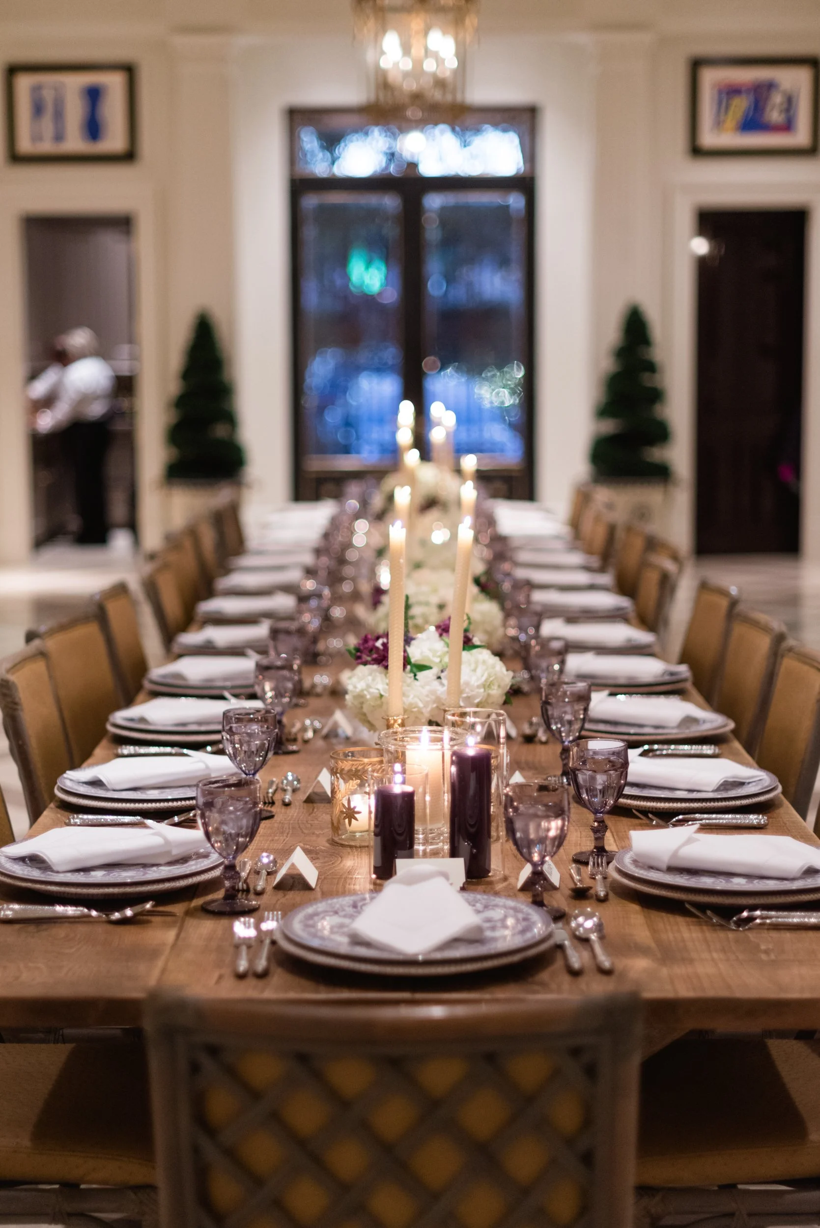 Elegant dining table set for a formal event with white plates, folded napkins, purple goblets, and tall candles as centerpieces in a decorated room.