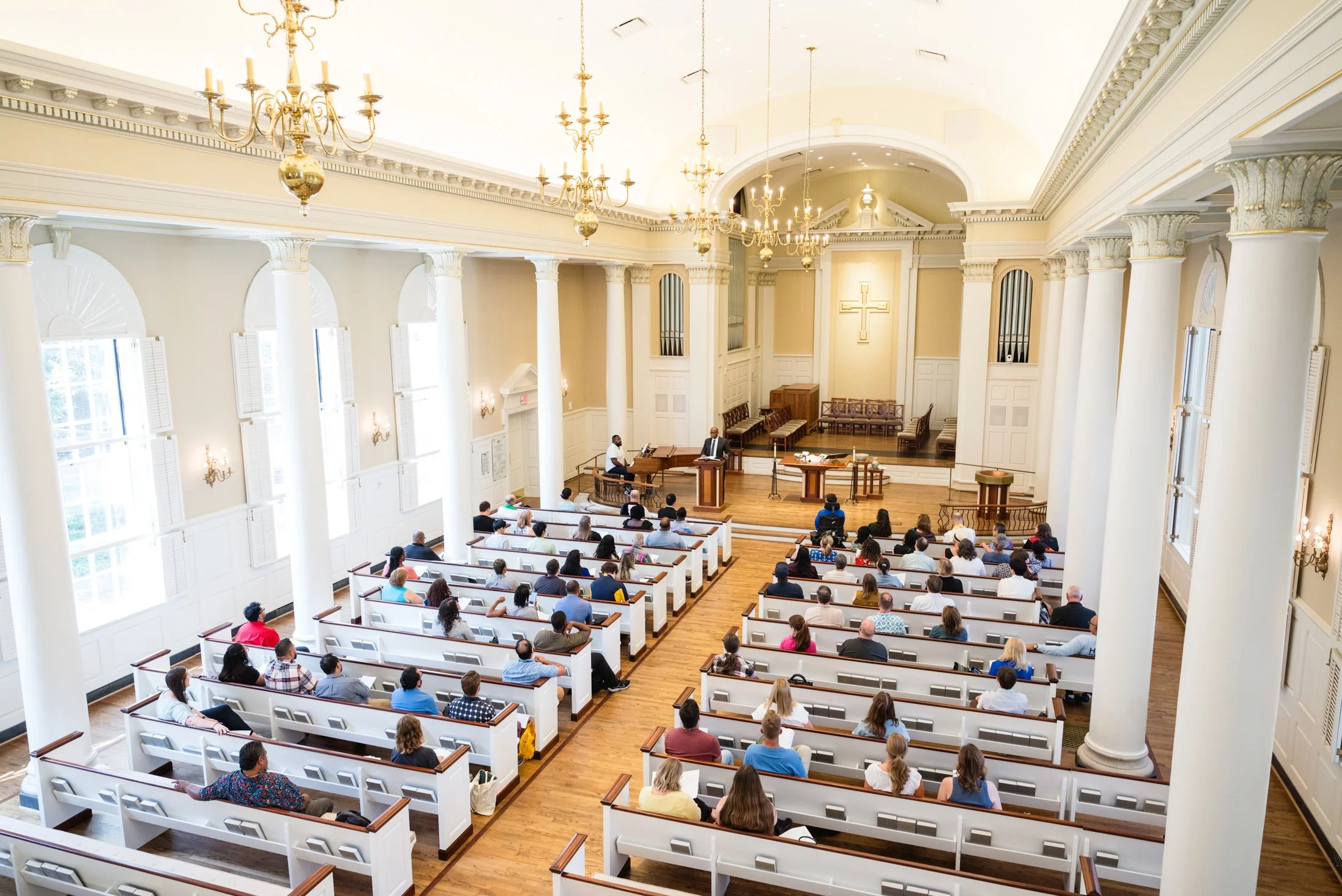 Interior of a church with white pews filled with people, a raised platform with pulpit, musical instruments, and a large cross on the wall behind the sanctuary.