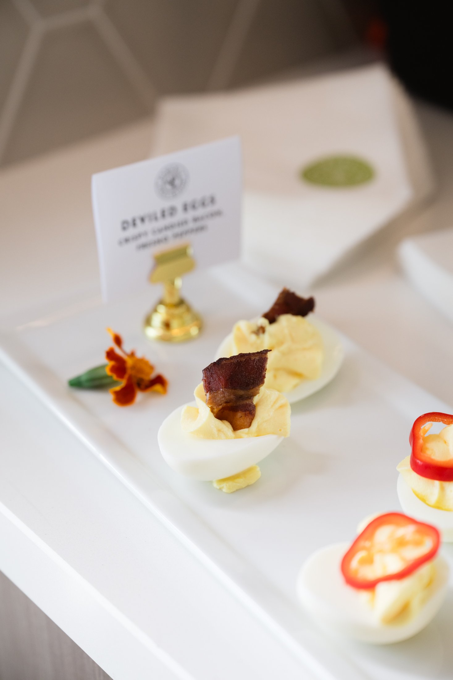 Deviled eggs garnished with bacon slices, placed on a white serving platter, with a small floral decoration and a sign indicating 'Deviled Eggs' in the background.