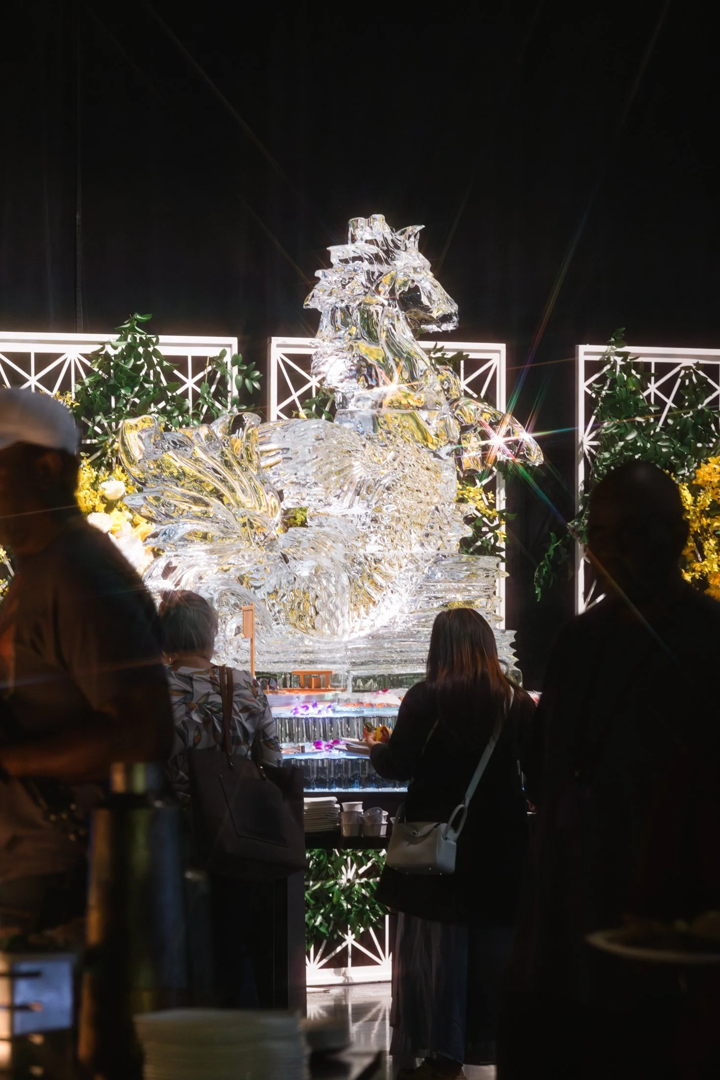 A large ice sculpture of a horse surrounded by plants and lighting, with people admiring it.
