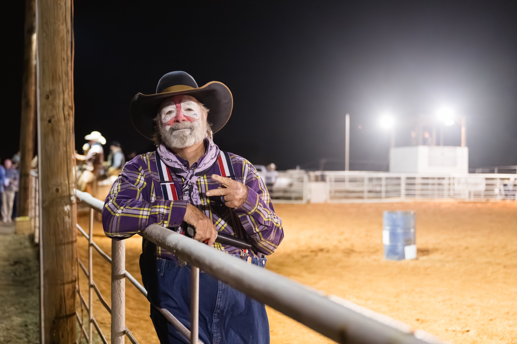 A man dressed as a clown or performer at a rodeo or fair, wearing face paint, a wide-brimmed hat, a plaid shirt, and suspenders, standing by a metal fence with his hands resting on it, making a peace sign and looking at the camera at night with brigh