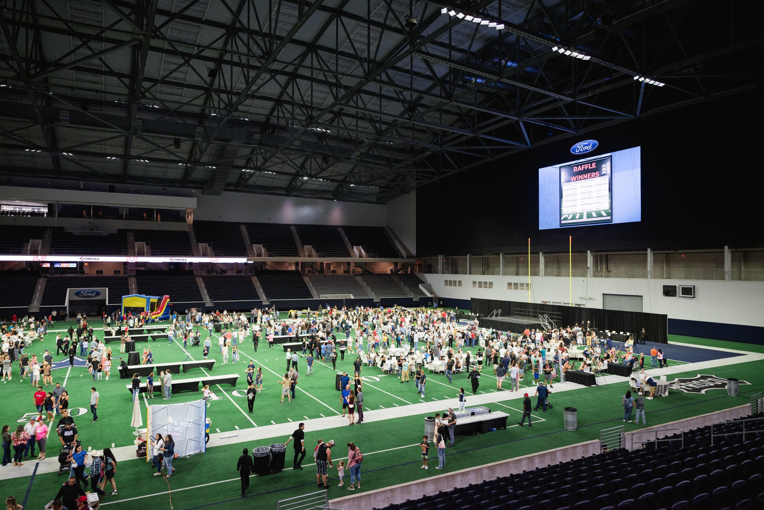 An indoor stadium filled with people attending an event on a turf field with inflatable attractions, tables, and chairs. A large screen displays a raffle winner announcement, and the seating area is mostly empty.