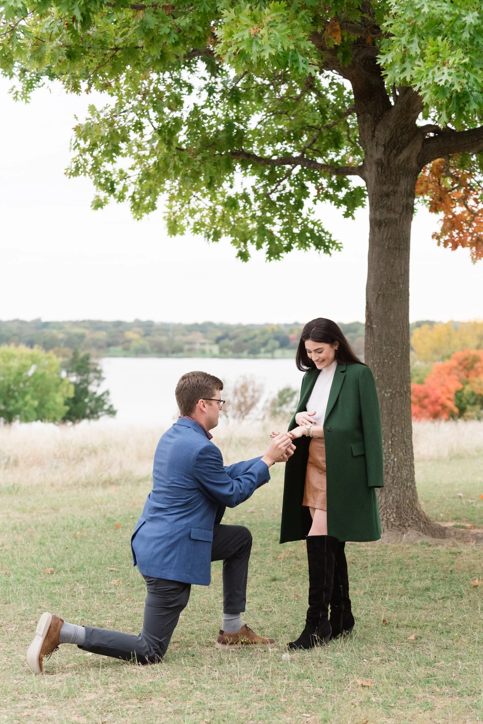 A man proposing marriage to a woman outdoors under a tree by a lake during fall, with the man on one knee holding her hand and the woman smiling.