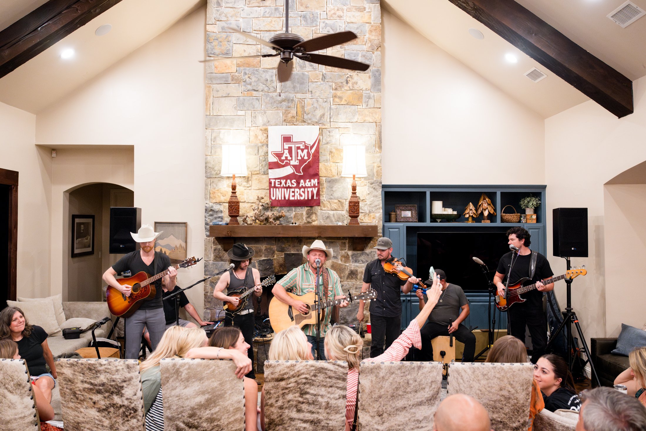 A band performing on stage in a cozy living room with a stone fireplace and a Texas A&M University banner above it. The band includes guitarists, a violinist, and a drummer, with an audience of children and adults enjoying the show.