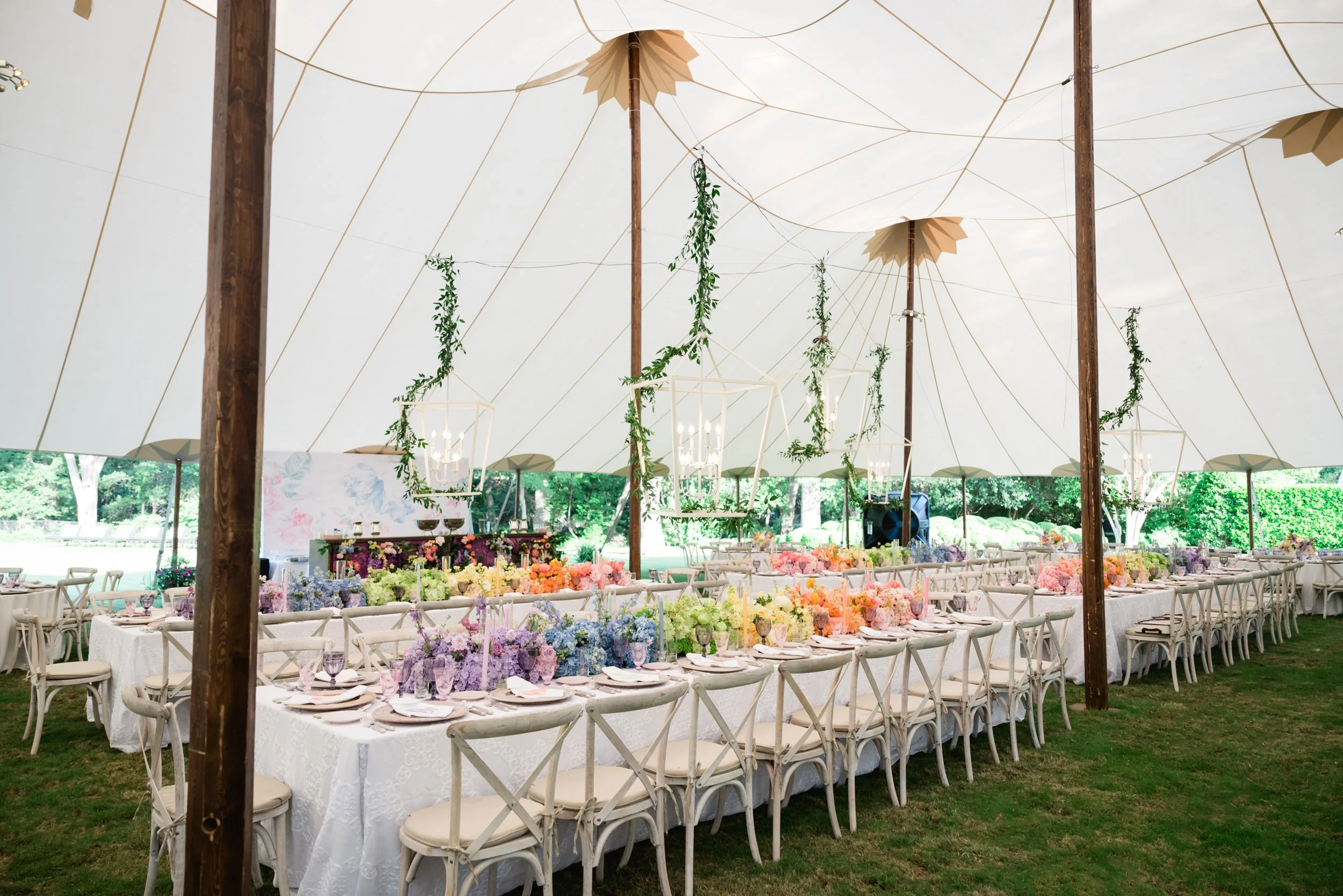 Wedding reception under a large white tent with long tables decorated with pastel-colored floral centerpieces, white tablecloths, and chairs. Greenery and hanging lanterns with candles are suspended from the tent ceiling.